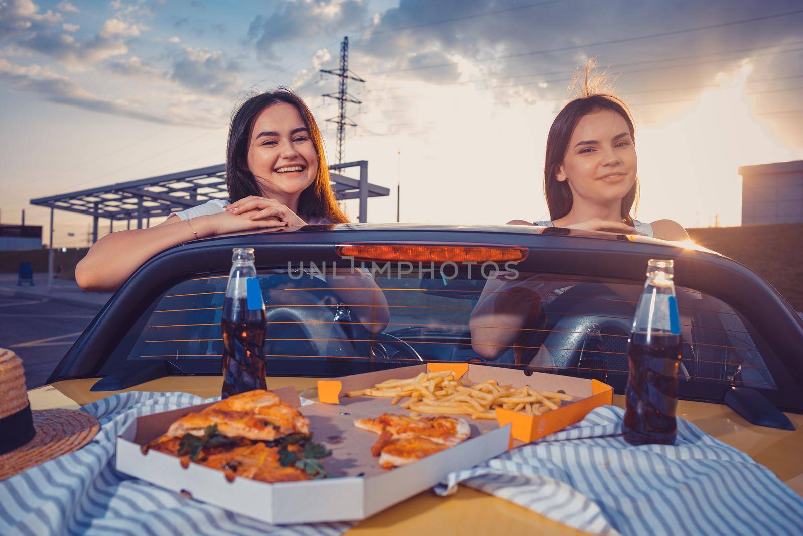 Young women in casual clothes are smiling, posing in yellow car cabriolet with french fries, pizza and soda in glass bottles on its trunk. Fast food. Summer evening. Close up, copy space, mock up
