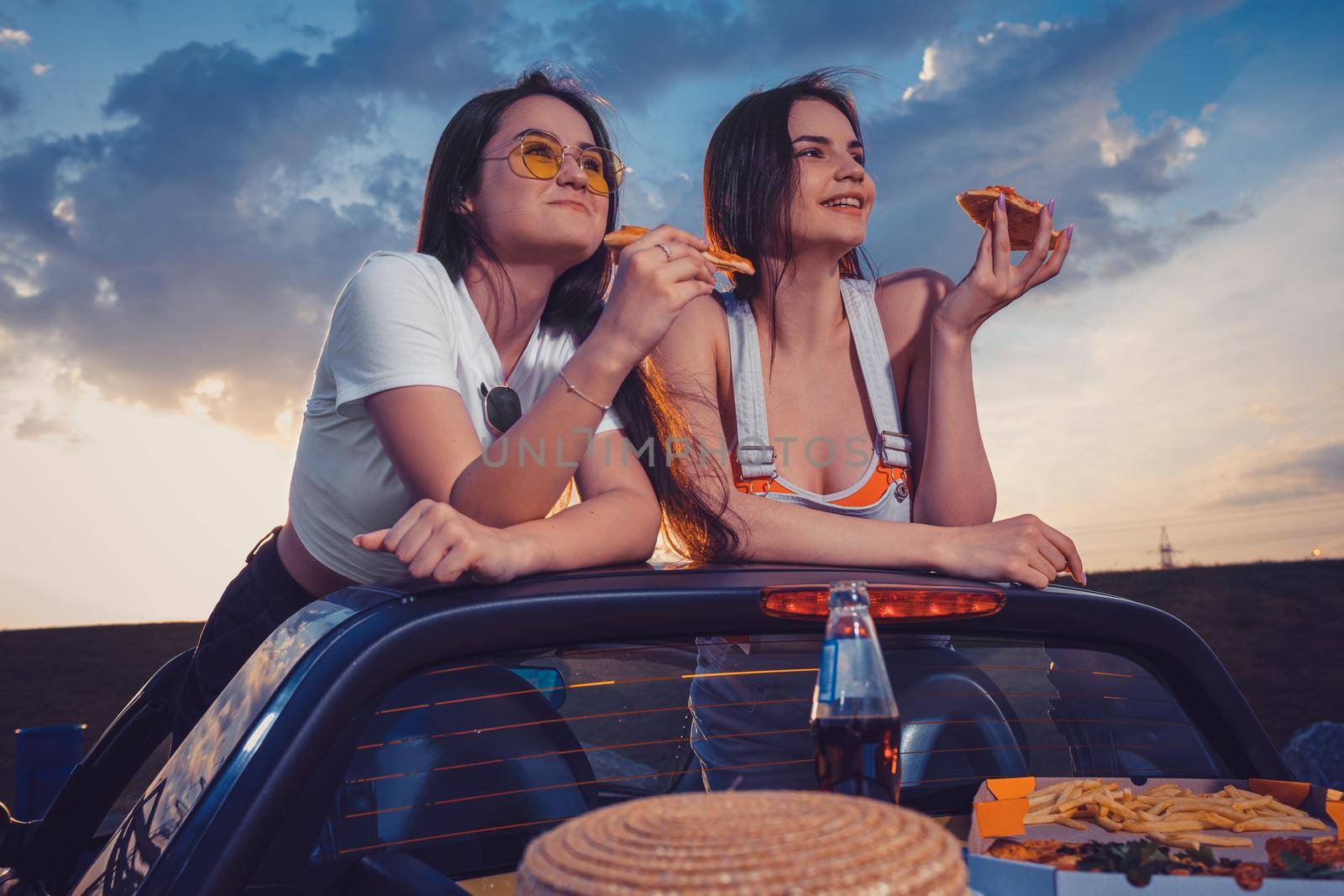 Two young women in casual outfit eating pizza, smiling, posing in yellow car cabrio with french fries, hat and soda in glass bottle on trunk. Fast food. Summer sunset, cloudy sky. Close up, mock up