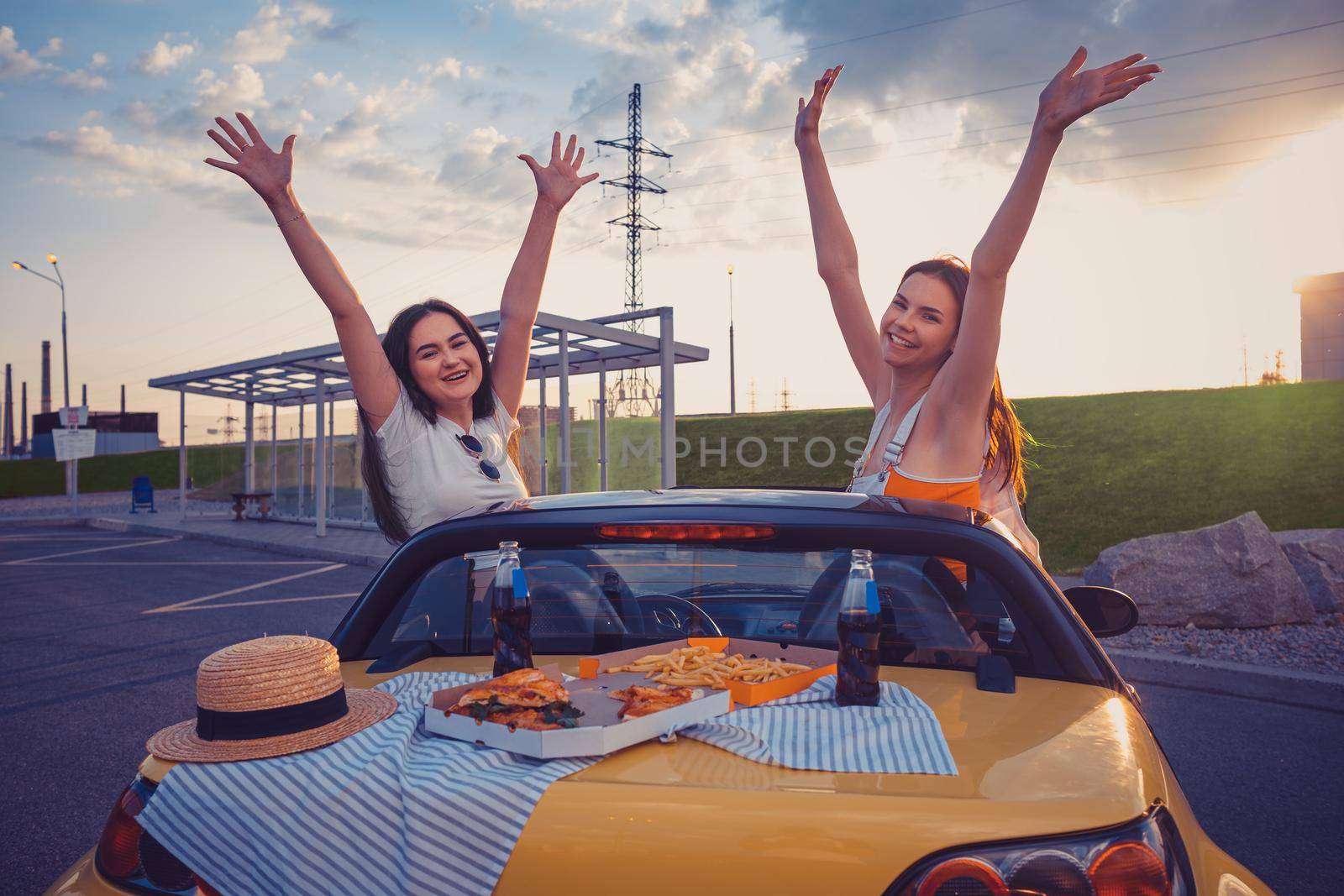 Young ladies in casual outfit are smiling, raised up hands, posing in yellow car with french fries, pizza and soda on its trunk. Fast food. Summer evening, bus stop. Close up, copy space