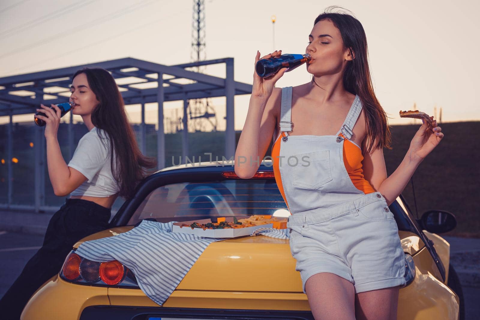 Attractive females in casual outfit are drinking carbonated beverage in glass bottles, eating pizza, posing near yellow car on parking lot. Fast food. Summer evening. Close up, copy space, mock up