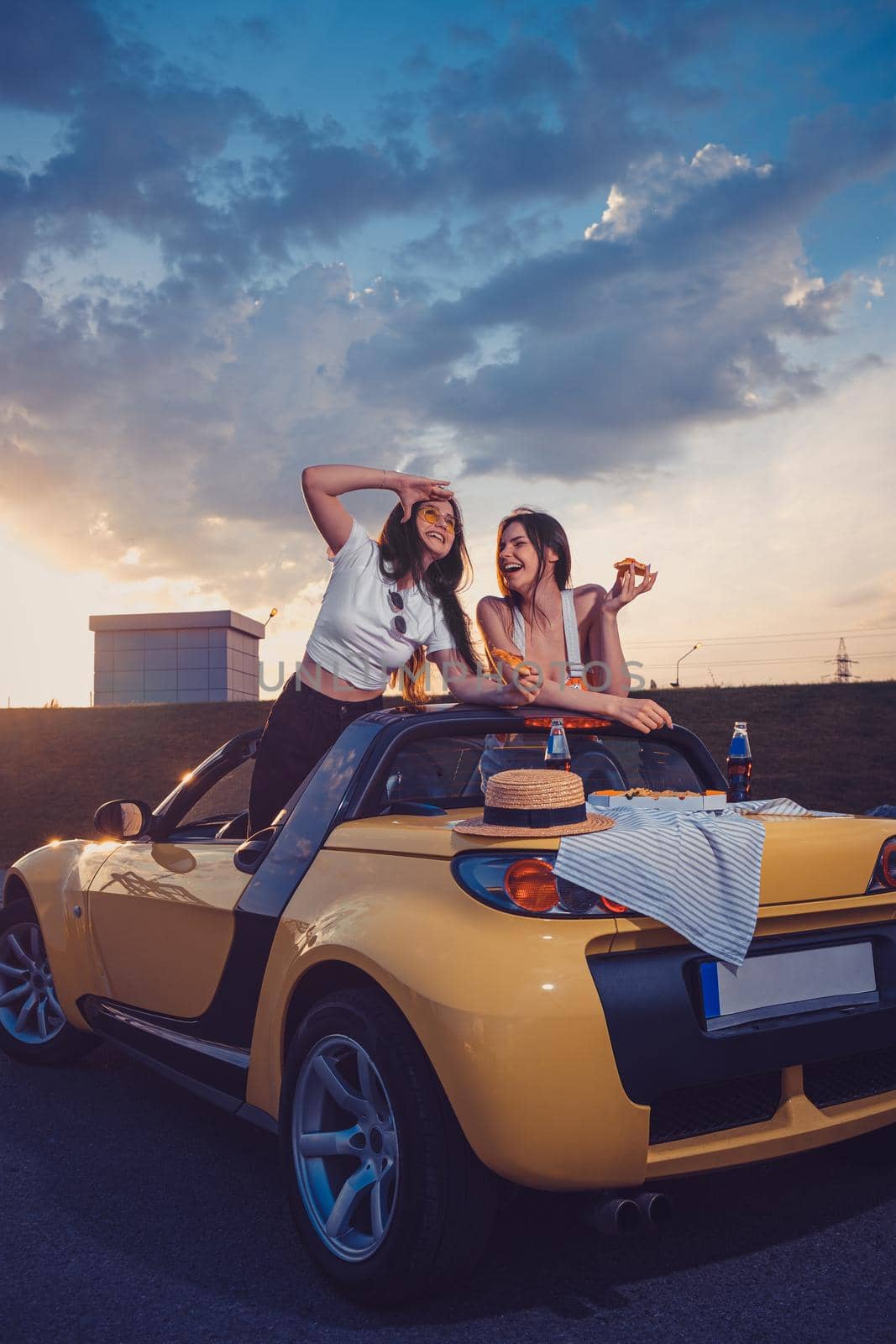 Two young models eating pizza, laughing, posing in yellow car cabriolet with hat and soda in glass bottles on its trunk. Fast food. Copy space by nazarovsergey