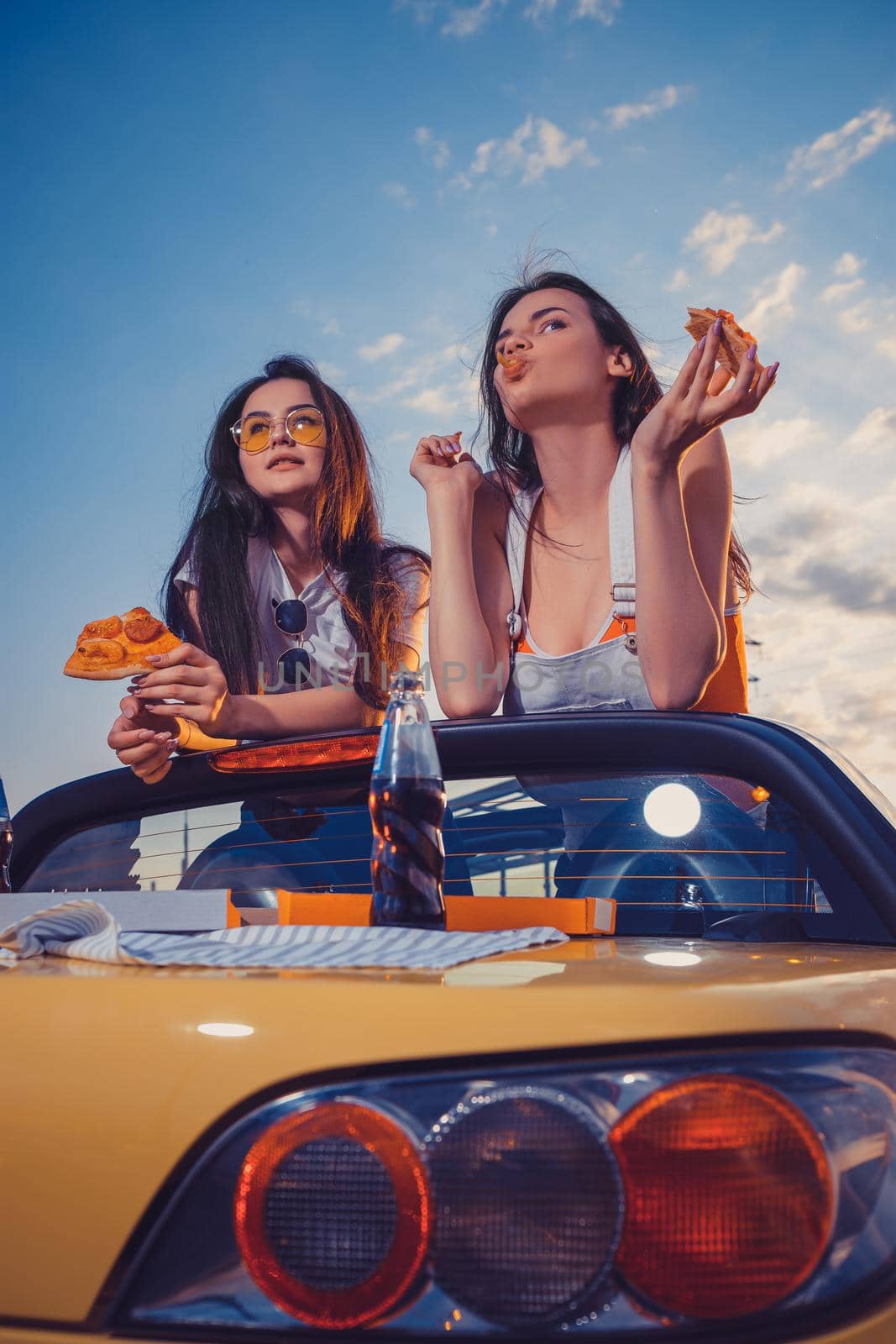 Two lovely females eating pizza while posing in yellow car cabriolet with soda in glass bottle on its trunk. Fast food. Close up, copy space by nazarovsergey