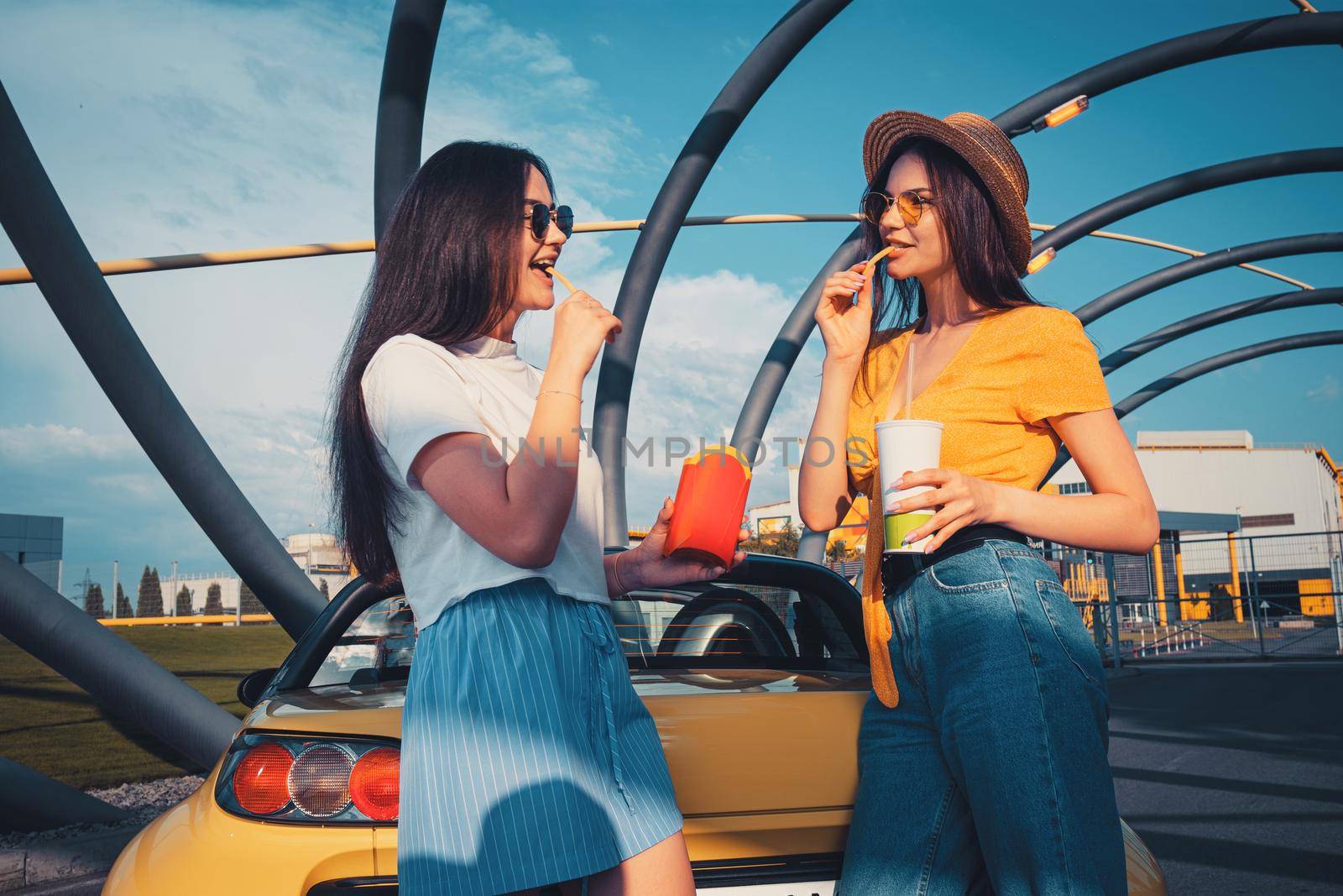Young females in casual outfit are smiling, eating french fries and holding beverage in paper cup while posing near yellow car roadster. Fast food. Sunny day, industrial zone. Close up, copy space