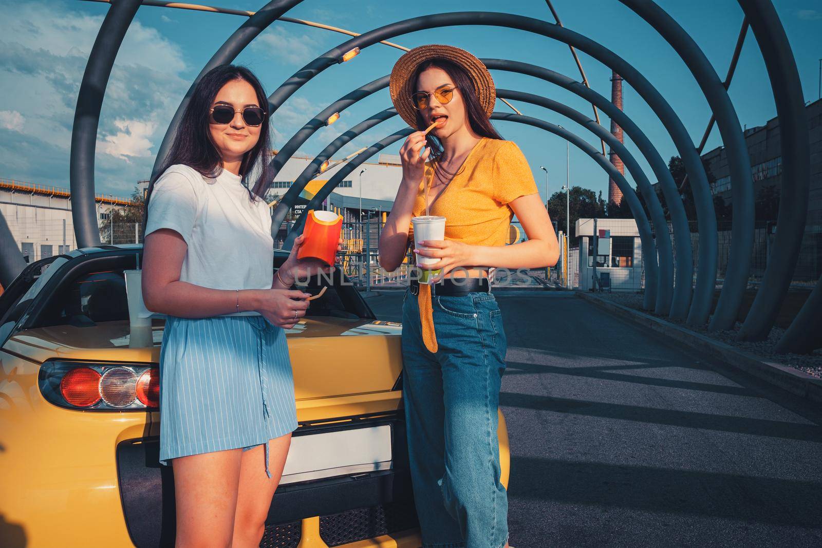 Young girls in casual clothes are smiling, enjoying french fries from paper package and beverages in paper cups, posing near yellow car. Fast food. Sunny day, industrial zone. Close up, copy space