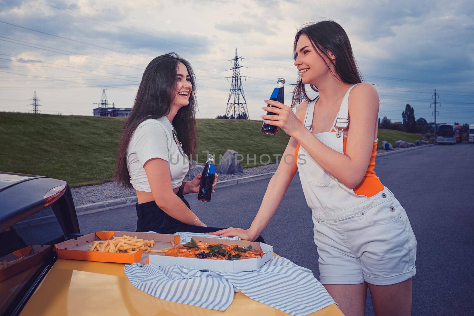 Young girls in casual clothes are smiling, enjoying soda in glass bottles, posing leaning on trunk of yellow car with pizza and french fries on it. Fast food. Parking lot. Close up, copy space