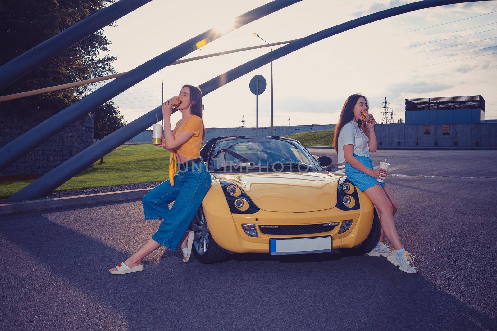 Young models in casual clothes are eating hamburgers and holding beverages in paper cups while leaning on the hood of yellow car roadster. Fast food. Sunny day, industrial zone. Copy space, mock up