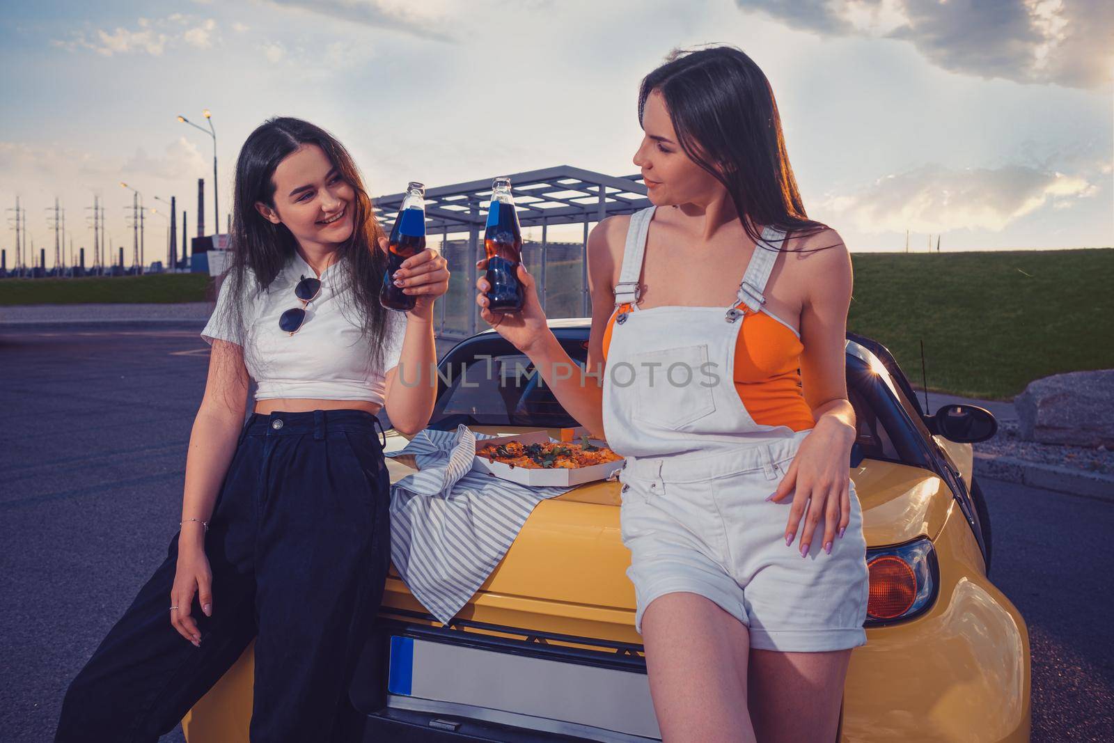 Gorgeous females are cheering with soda in glass bottles while leaning on trunk of yellow car with pizza on it. Fast food. Close up, copy space by nazarovsergey