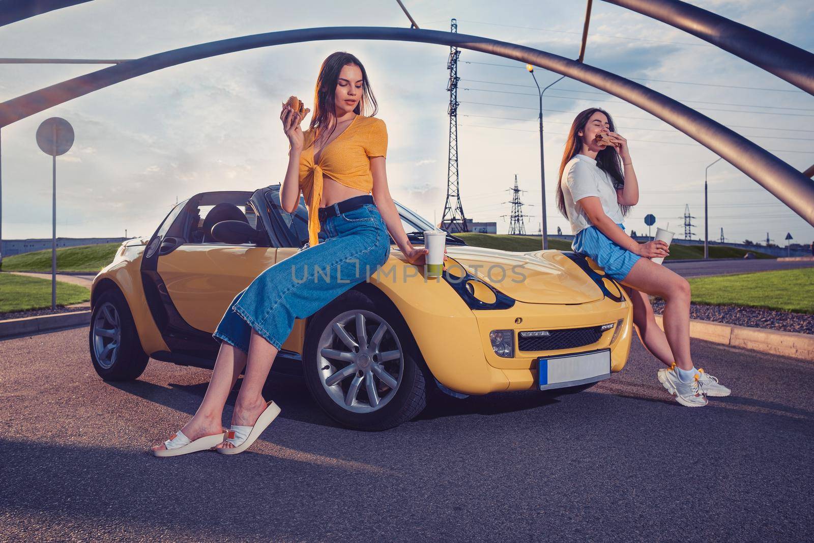 Attractive women in casual clothes are eating hamburgers and holding drinks in paper cups while leaning on the hood of yellow car cabrio. Fast food. Sunny day, iron pipes arch. Copy space, mock up
