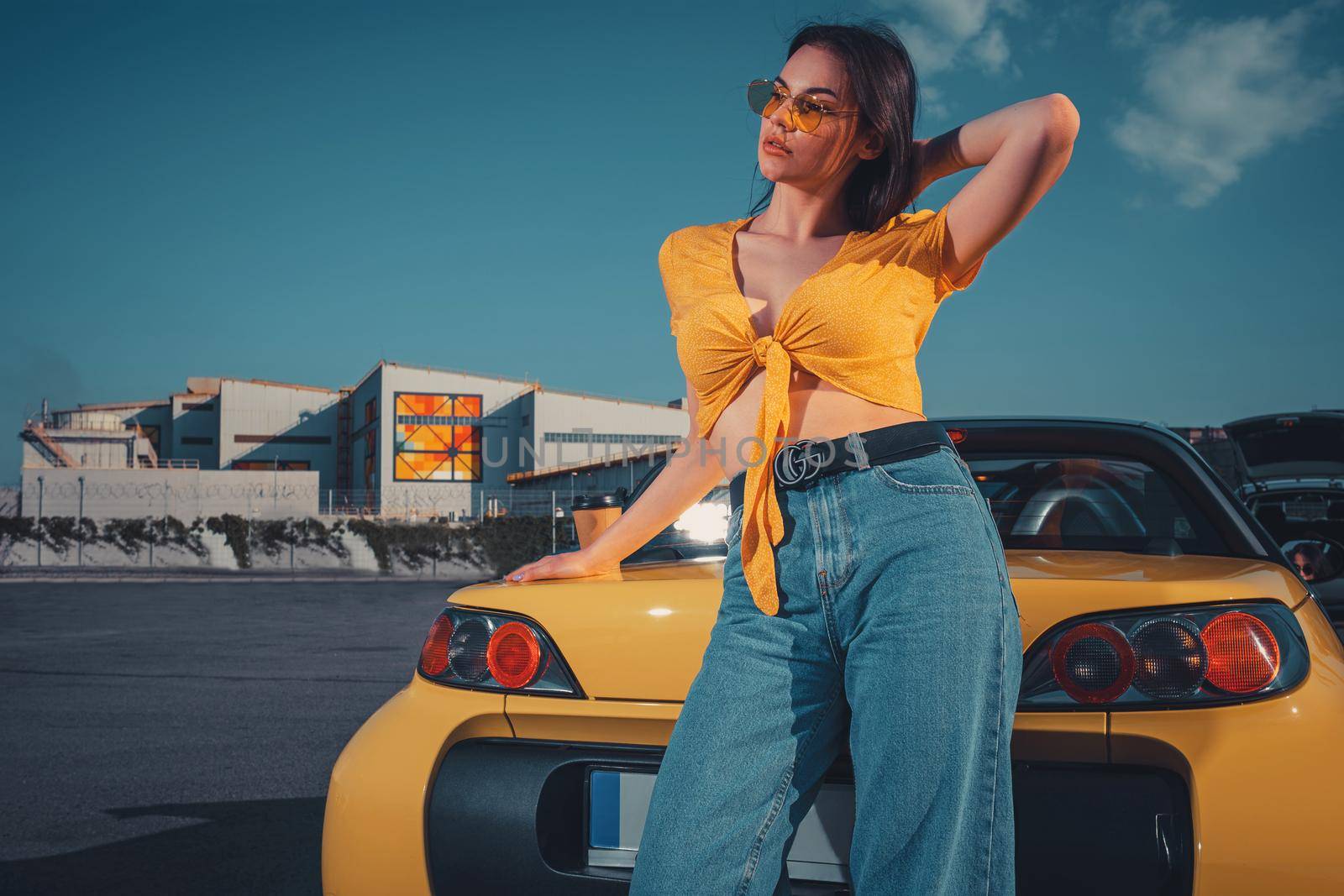 Young brunette woman in blue jeans, orange top and sunglasses is posing near yellow car cabriolet with paper cup of coffee on trunk. Summer sunny day at parking lot. Close up, copy space, mock up