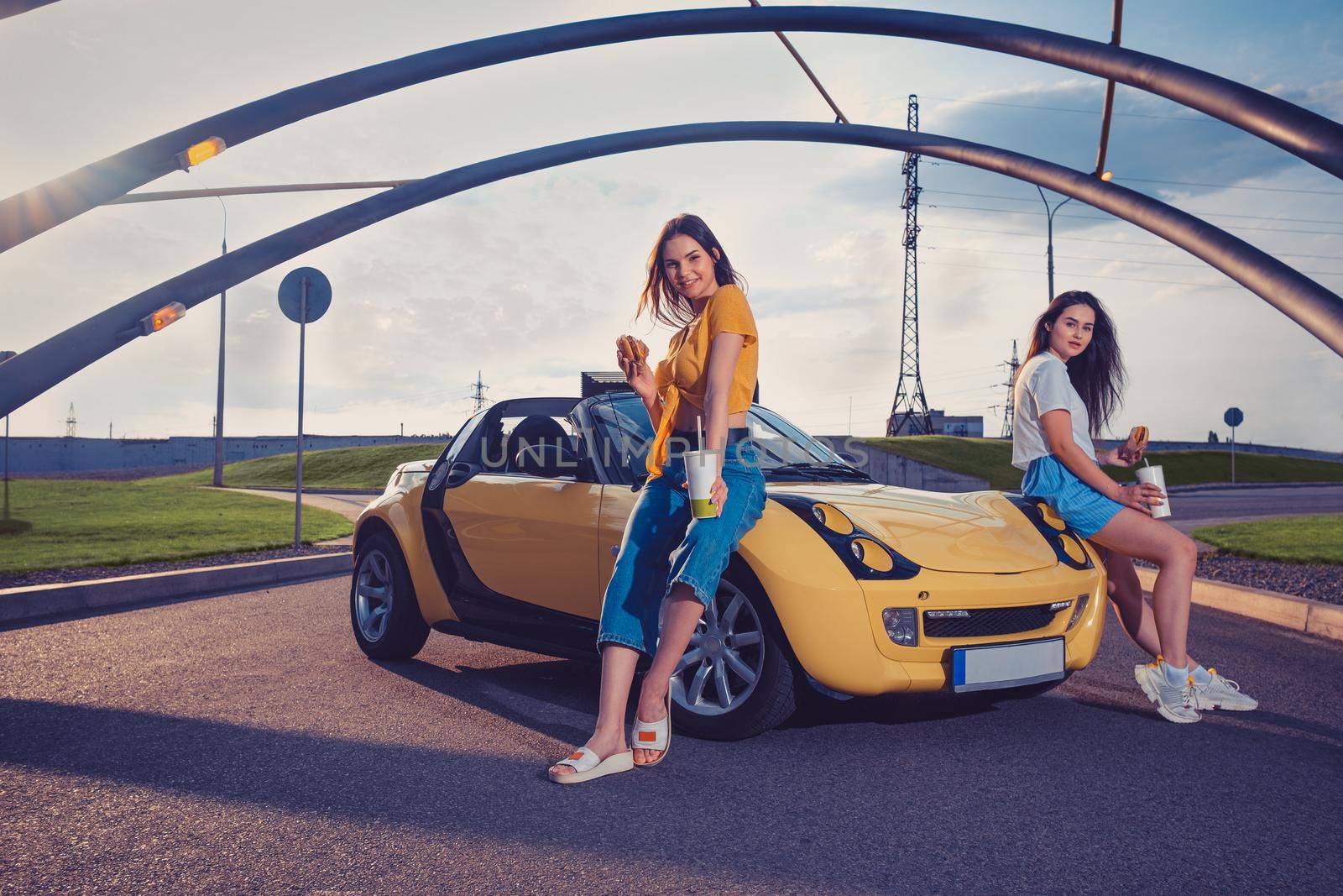Gorgeous ladies in casual clothes are smiling, holding hamburgers and beverages in paper cups while sitting on the hood of yellow car cabrio. Fast food. Sunny day, iron pipes arch. Copy space, mock up