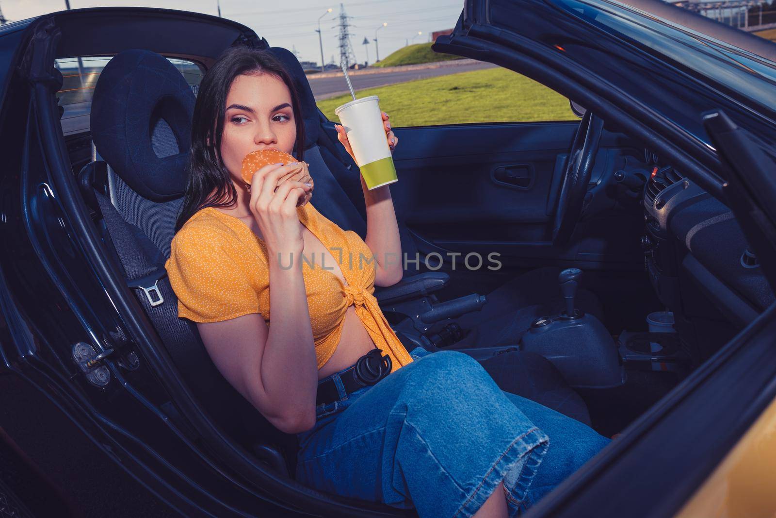 Attractive model in blue jeans and orange top is eating hamburger and holding beverage in paper cup while sitting in yellow car cabriolet. Fast food. Side view. Close up, copy space, mock up