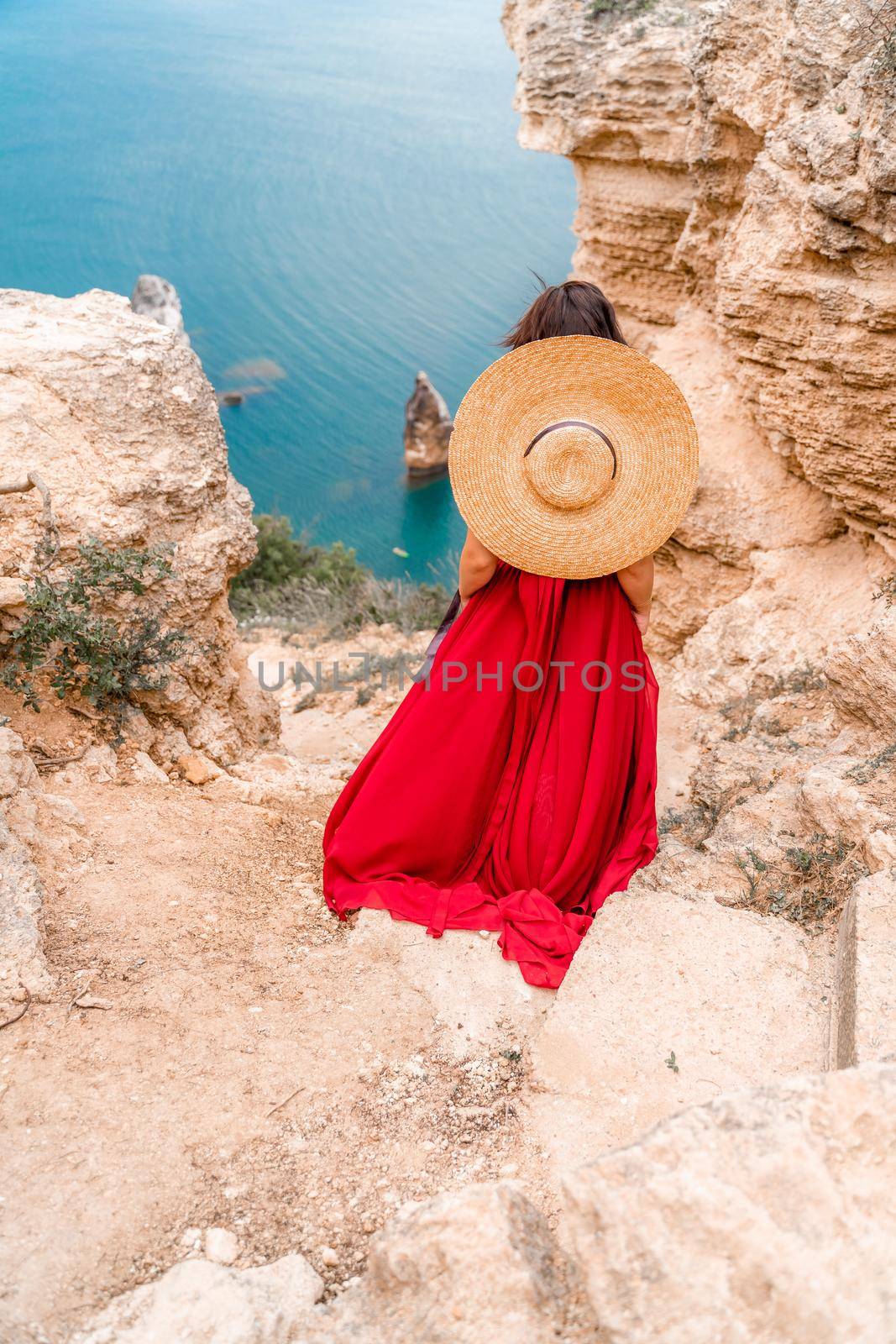 A girl with loose hair in a long red dress descends the stairs between the yellow rocks overlooking the sea. A rock can be seen in the sea. Sunny path on the sea from the rising sun by Matiunina