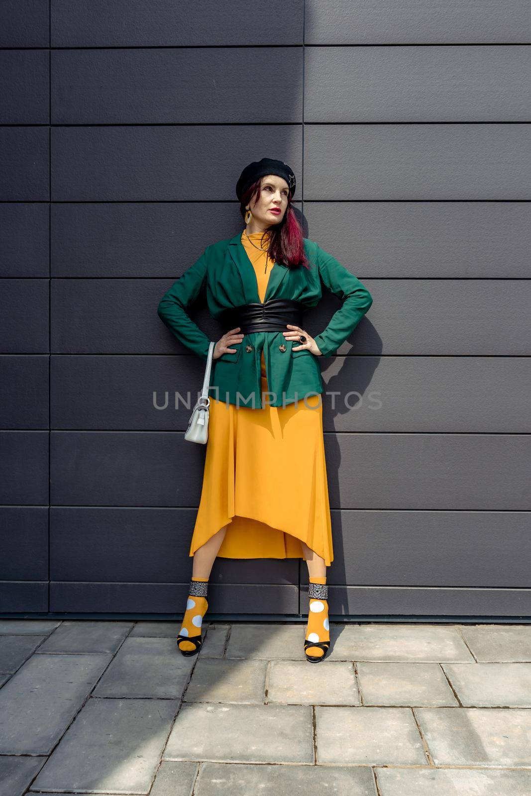 Young woman in bright clothes, yellow skirt and green jacket. Yellow socks in sandals, beret on the head, hair with the color of magenta. Caucasian female fashion model standing against gray wall background, open with copy space. by Matiunina