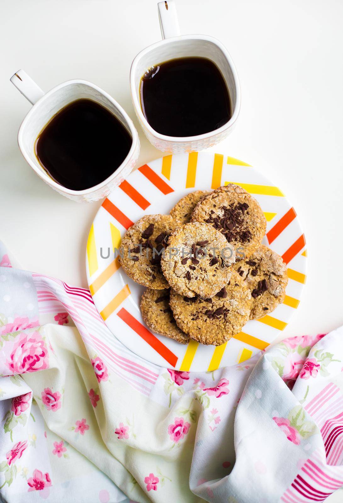 Oatmeal cookies with chocolate on a plate with bright cloth and two cups of coffee