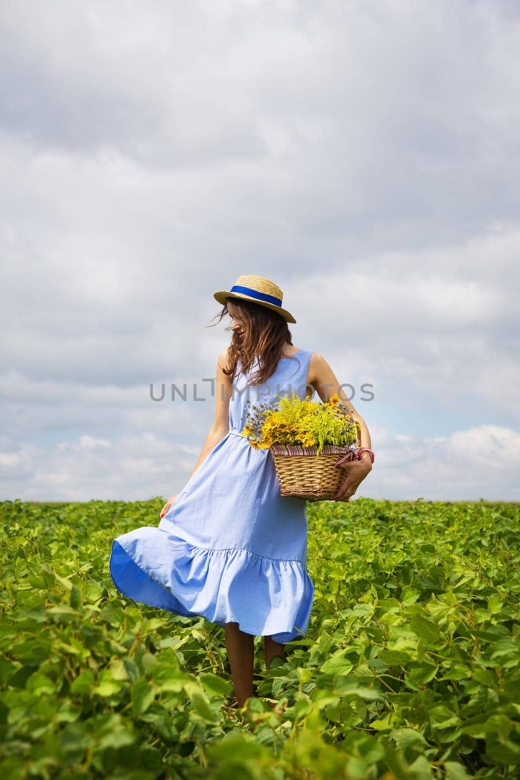 girl in a hat stands on a green field with a basket of flowers by sfinks