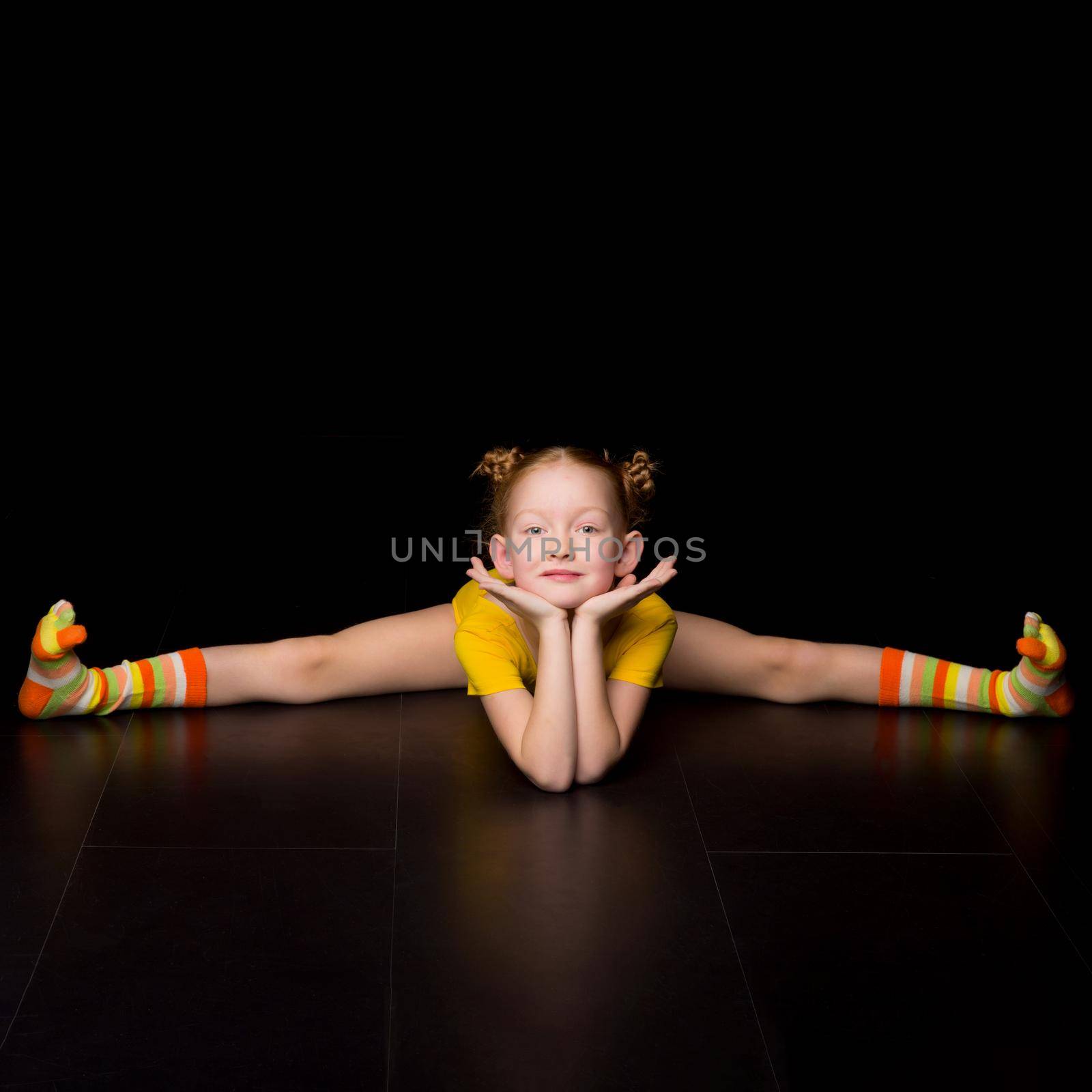Cute happy young girl gymnast doing cross splits. Happy adorable kid wearing yellow leotard performing rhythmic gymnastic exercise and smiling at camera against black background