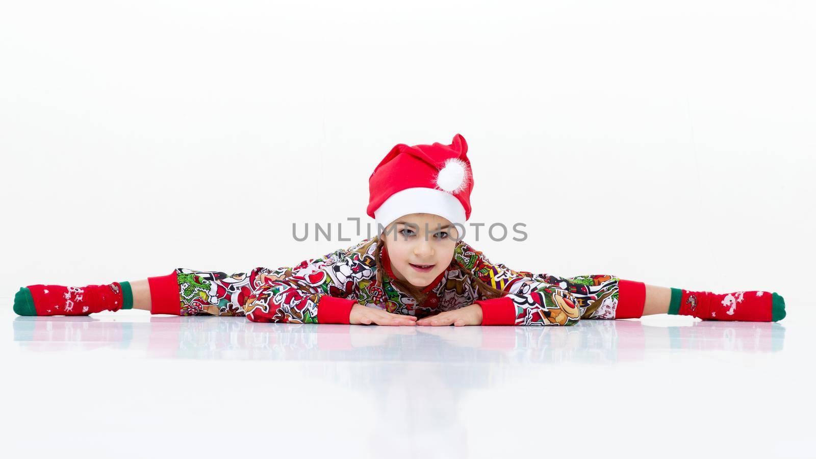 Happy flexible little girl gymnast doing cross splits. Cheerful girl wearing bright colorful overalls, socks and Santa Claus cap performing splits against isolated white background