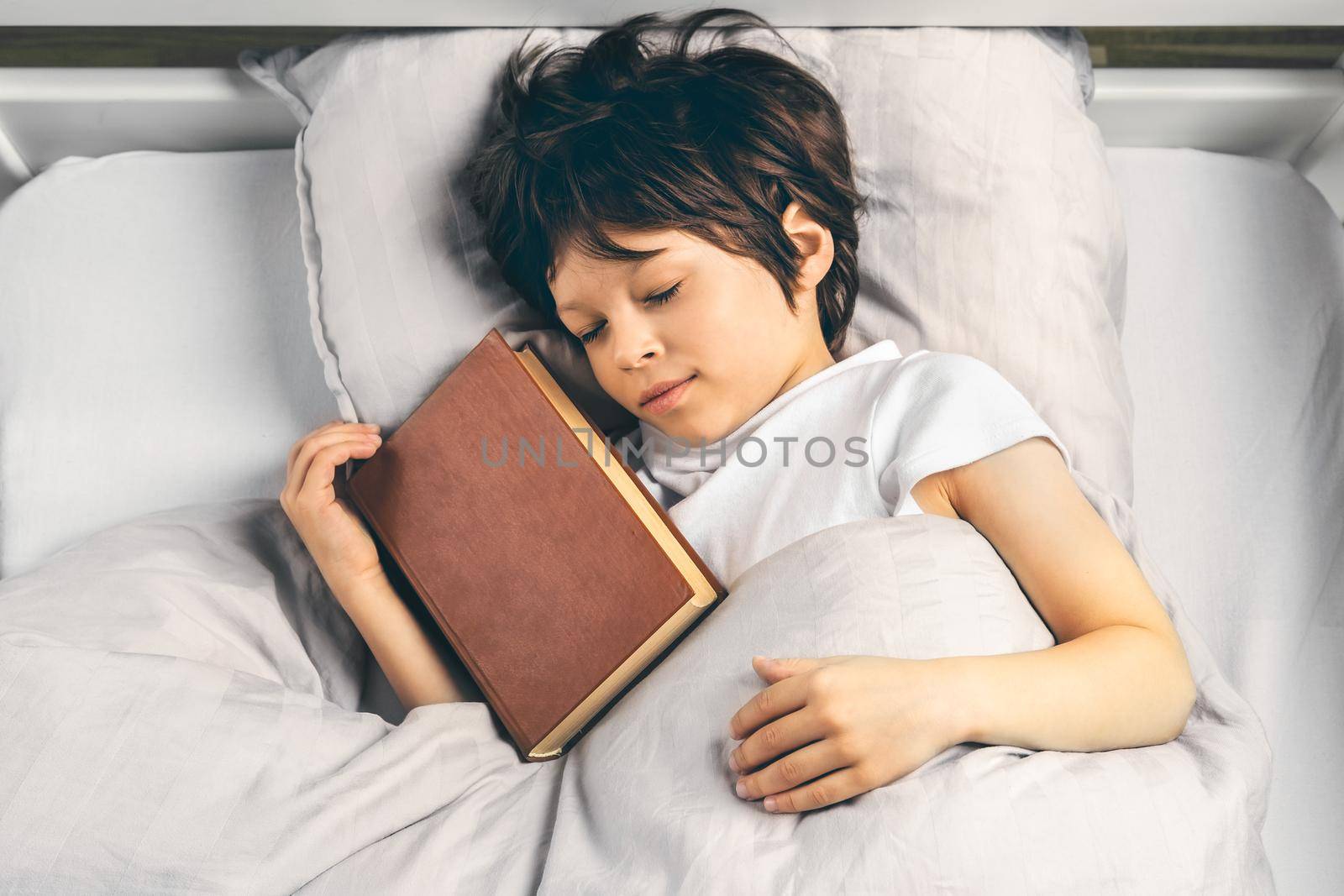 Little school age boy is sleeping in his bed beside a book he was reading earlier at night