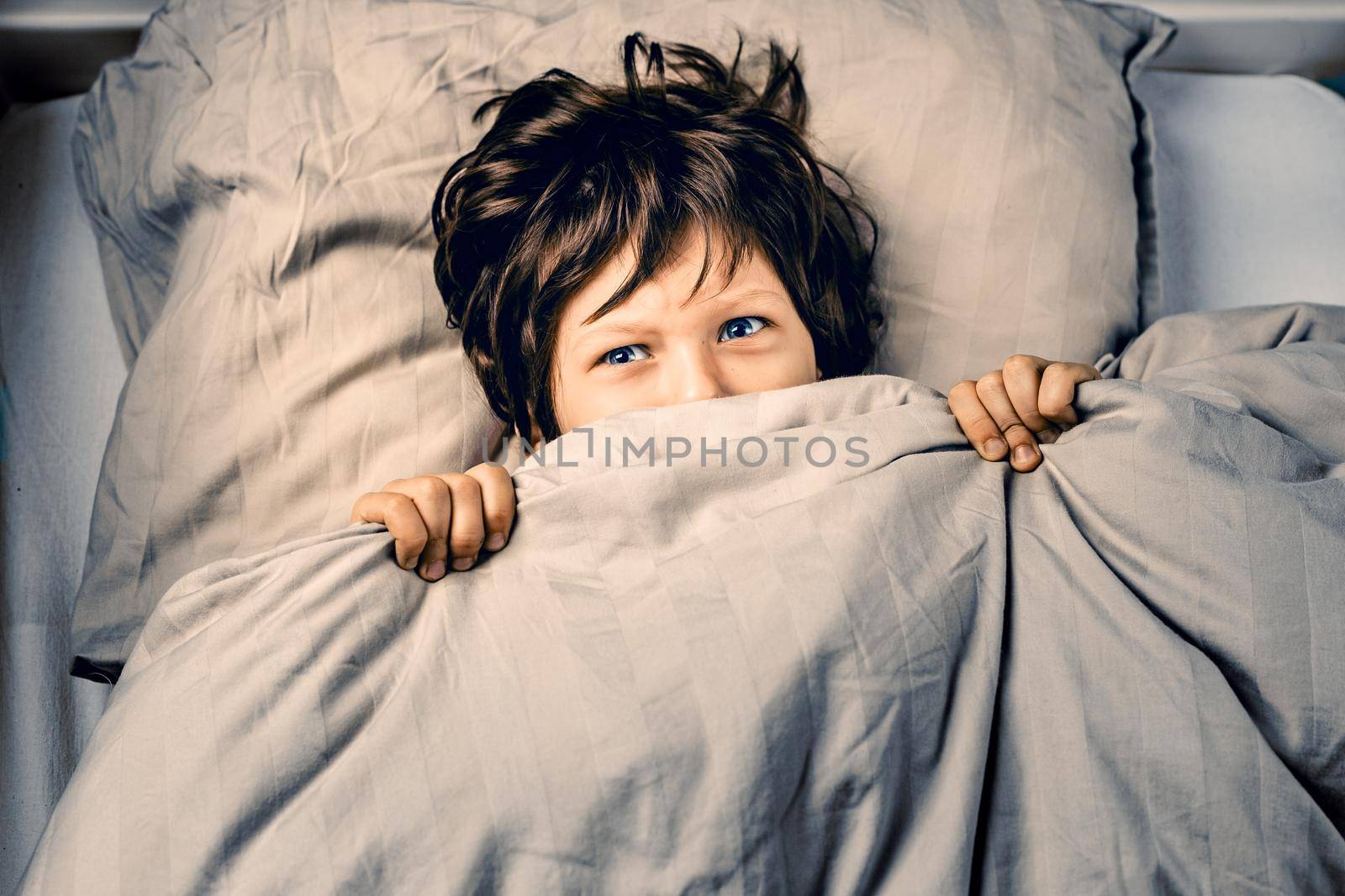 Little boy covering his face with a bed blanket is about to cry being scared of nightmares, ghosts or monsters in his bedroom. Child suffering from sleep disorder concept
