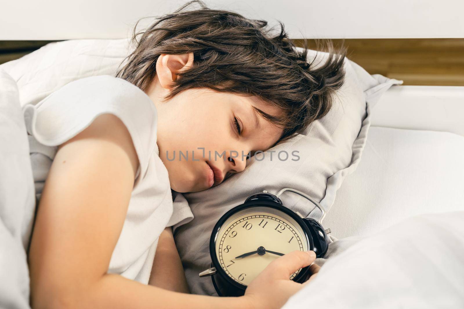 Adorable boy sleeping peacefully in his bed holding the traditional style alarm clock that shows fifteen minutes late the awakening time. Late for school or kindergarten, wake up time concept