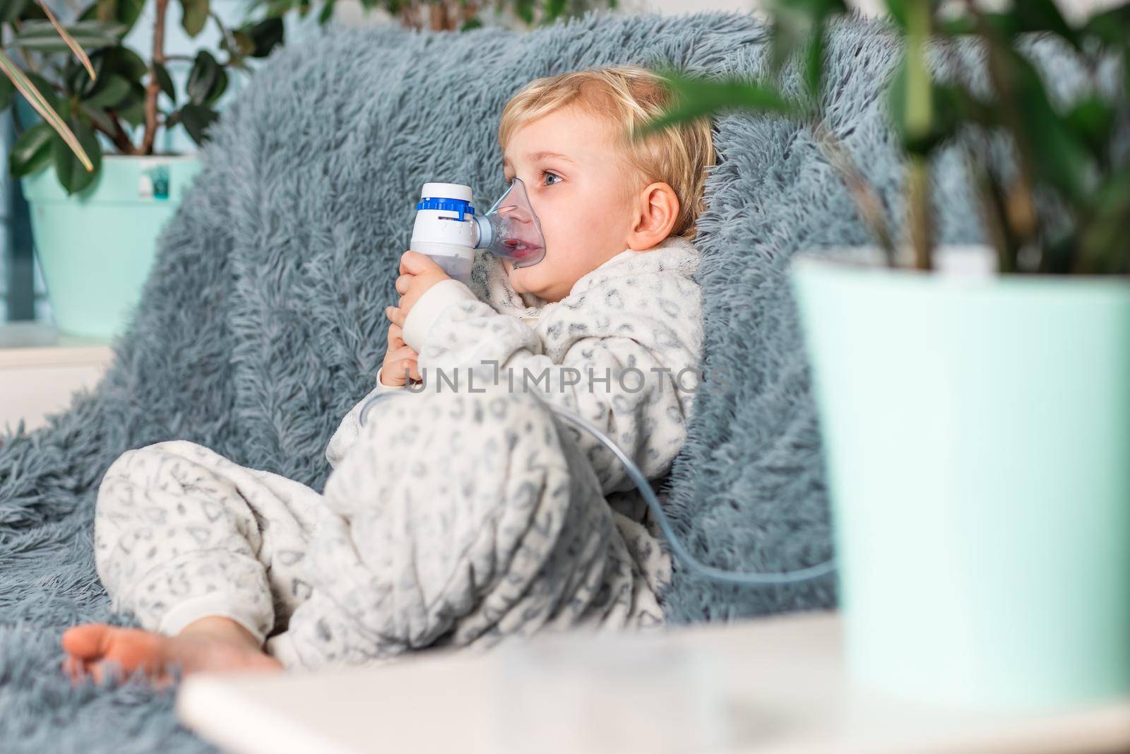 Cute baby boy makes inhalation with a nebulizer equipment. Sick child holding inhalator in hand and breathes through an inhaler at home by Len44ik