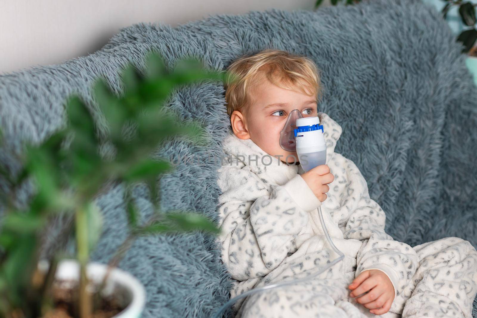 Cute baby boy makes inhalation with a nebulizer equipment. Sick child holding inhalator in hand and breathes through an inhaler at home. Physical therapy for cold, flu and bronchial asthma