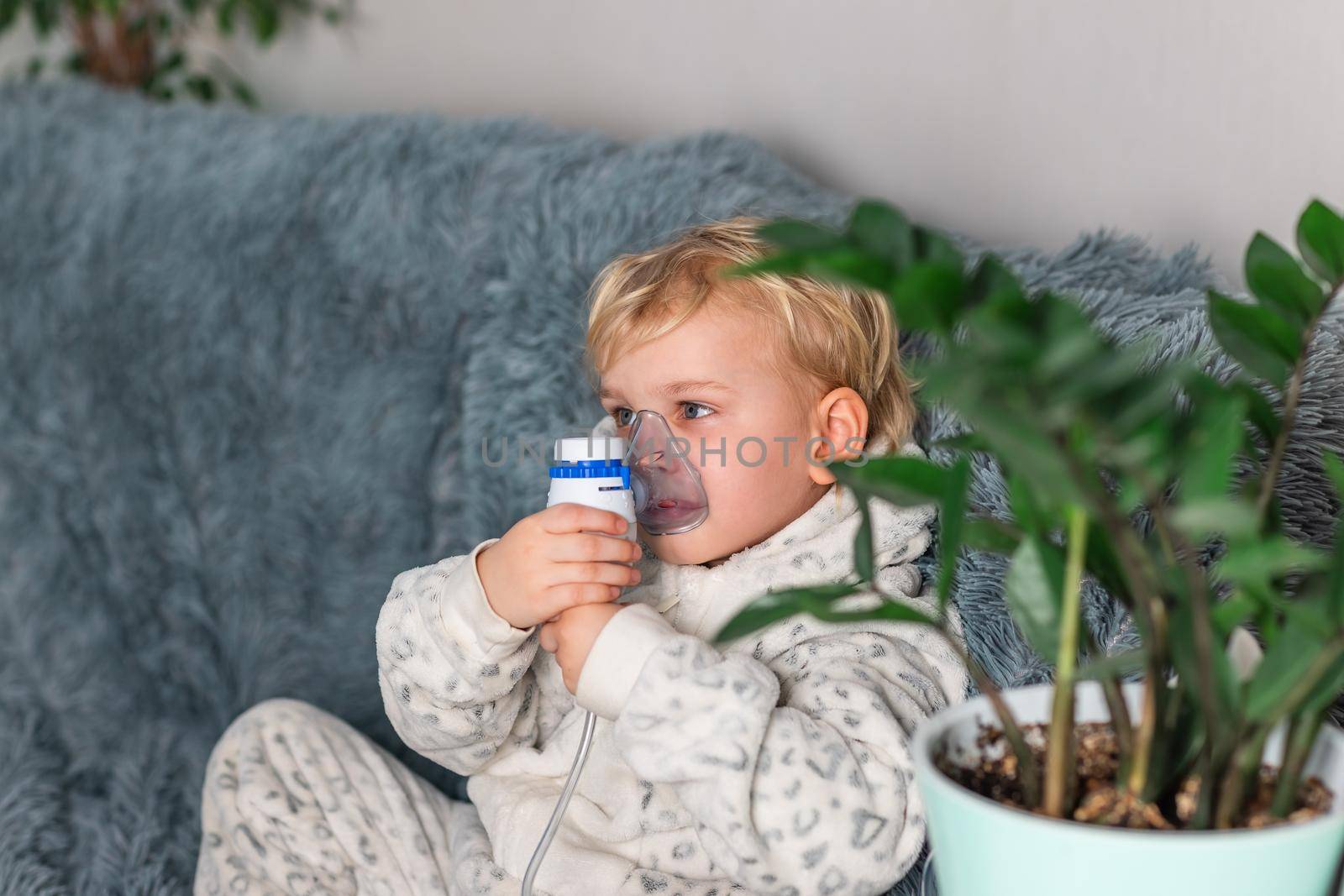 Cute baby boy makes inhalation with a nebulizer equipment. Sick child holding inhalator in hand and breathes through an inhaler at home. Physical therapy for cold, flu and bronchial asthma