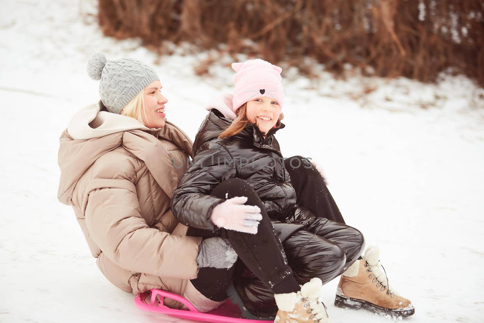 Happy mother with baby girl sitting on sledge and sledding down on snow from hill. Enjoying white winter day at park. Spending time together in weekend