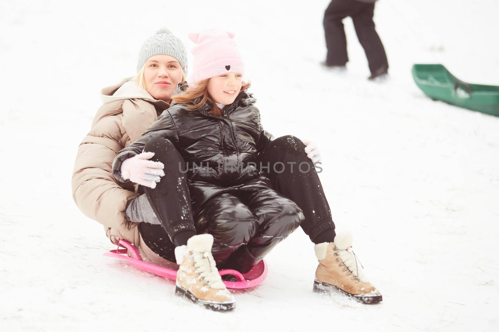 Happy mother with baby girl sitting on sledge and sledding down on snow from hill. Enjoying white winter day at park. Spending time together in weekend