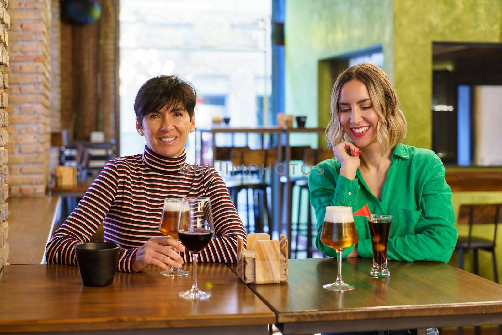 Happy adult women in casual clothes smiling and enjoying alcohol drinks while sitting at table during friends meeting on weekend day in pub