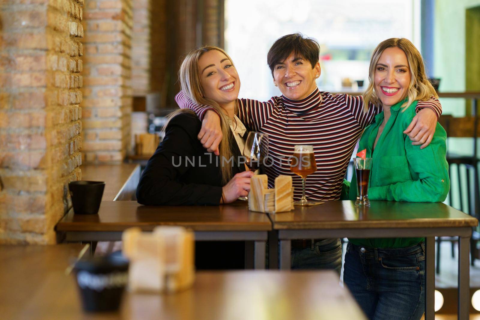Cheerful adult female with short dark hair looking at camera with smile and embracing girlfriends behind table with alcohol during meeting in pub