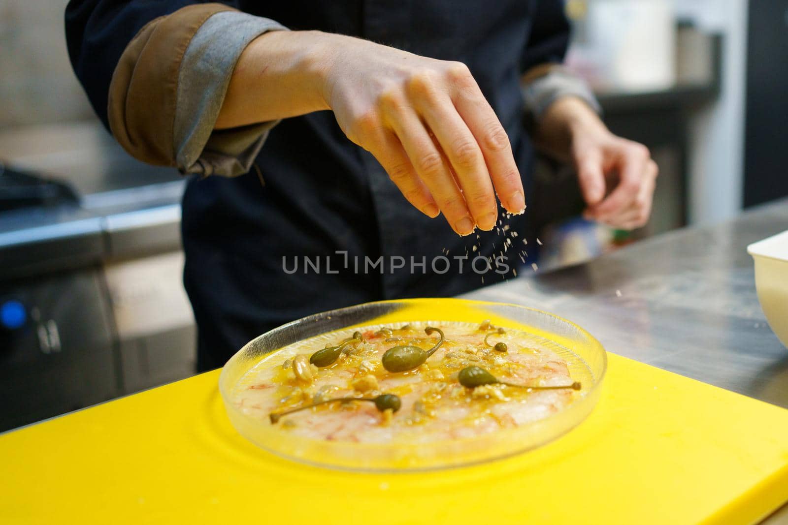Soft focus of anonymous cook scattering salt over plate of prawn carpaccio with capers during work in restaurant kitchen