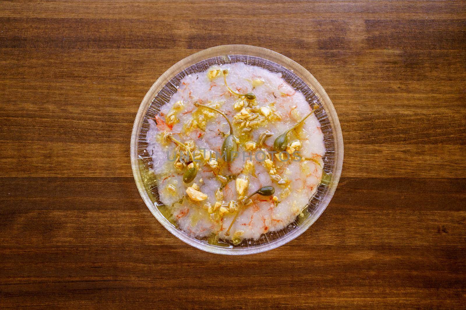 Top view of delicious dish with prawn carpaccio garnished with capers and nut sauce served on plate on wooden restaurant kitchen