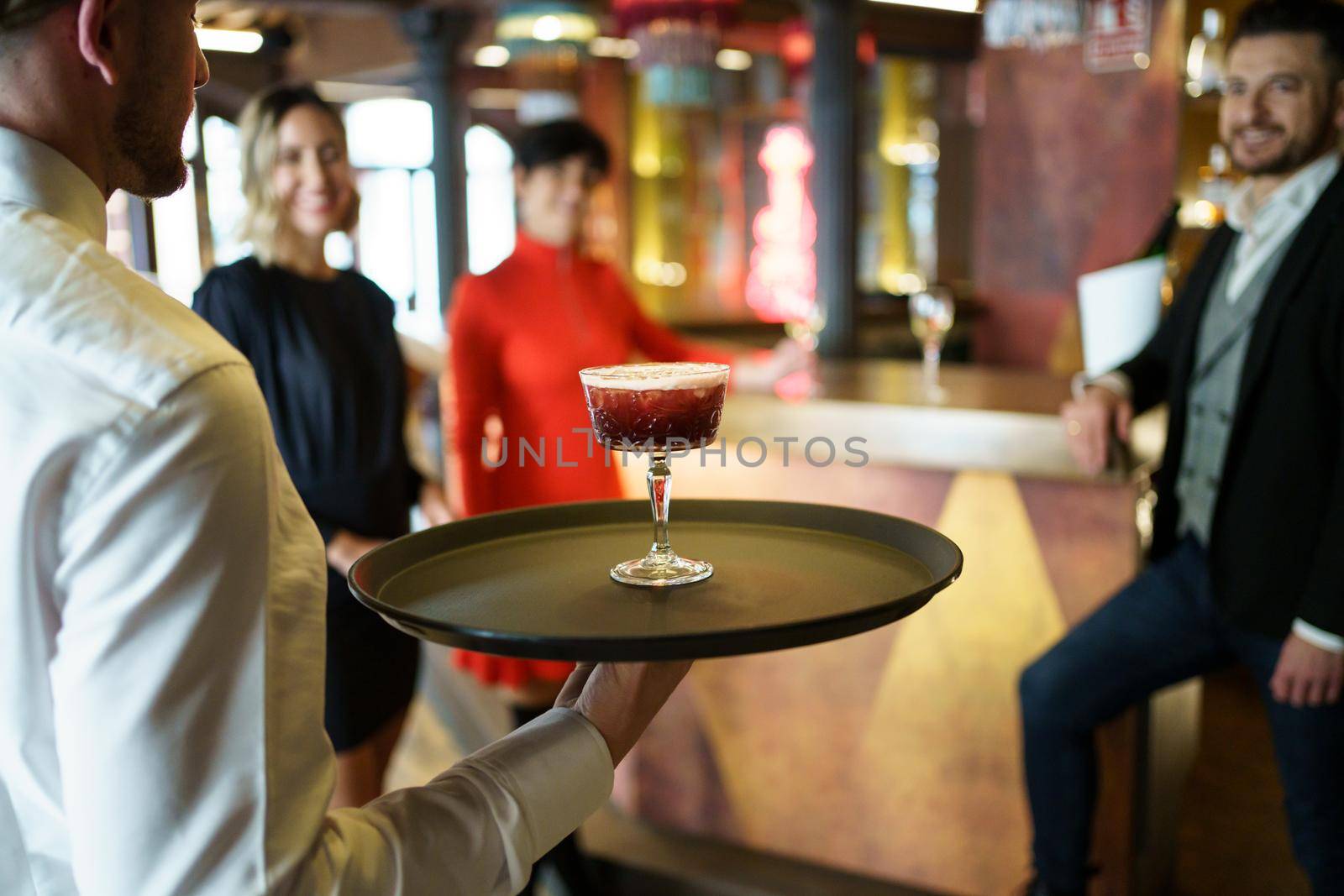 Back view of male bartender serving red cocktail on tray for company of elegant guests celebrating event in luxury restaurant