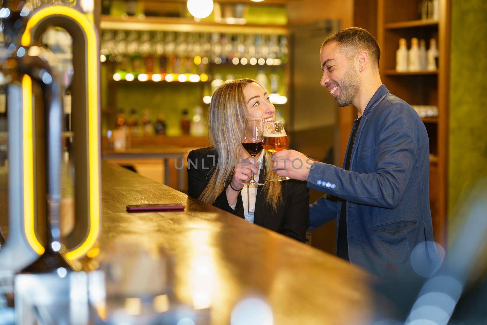 Happy diverse girlfriend and boyfriend looking at each other with smile and clinking glasses of alcohol drinks near counter during date in bar