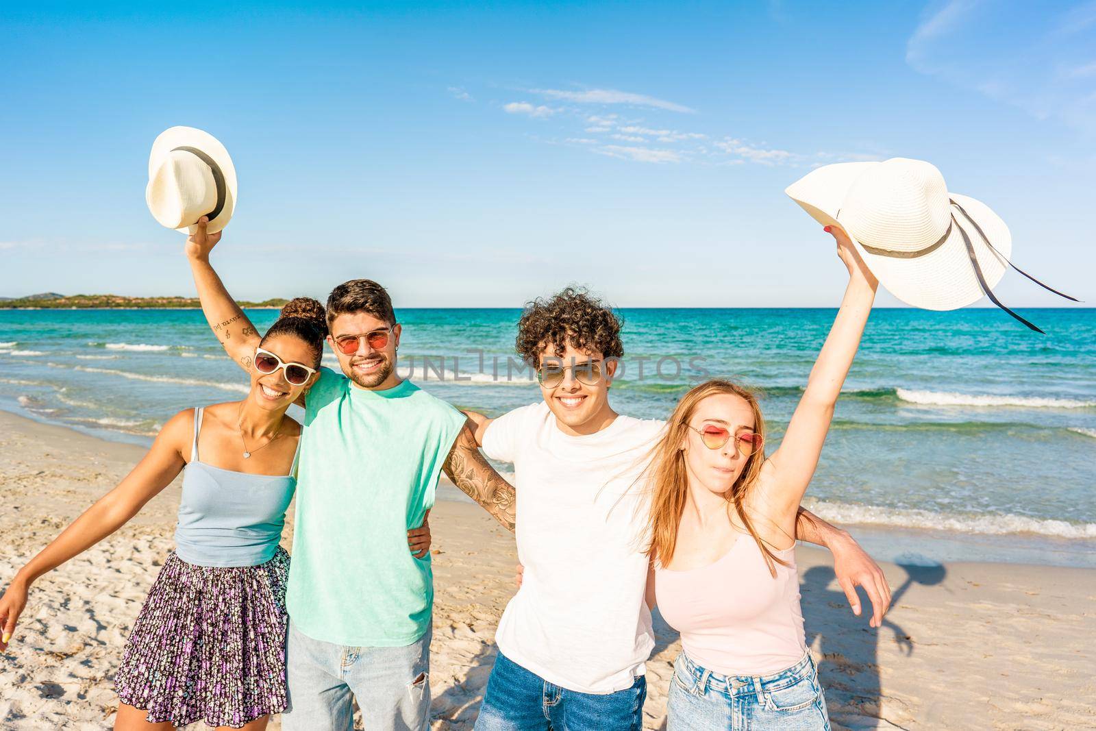 Group of gen z friends embracing to each other looking at camera on the seashore in tropical ocean resort. Happy students enjoying sea vacations together. Carefree people posing for photography
