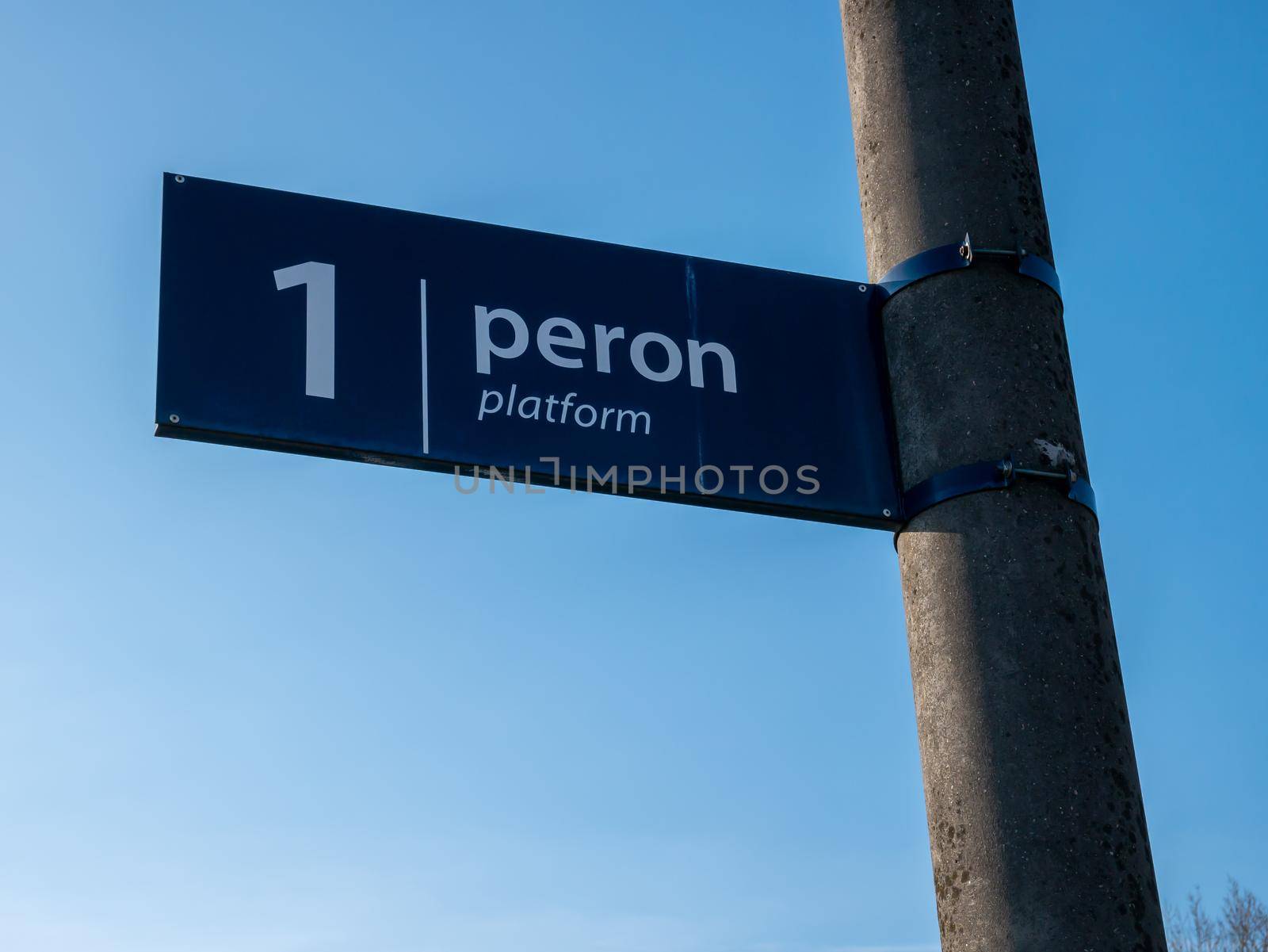 White and blue information sign on a train station in Poland with text Peron 1 in Polish and Platform 1 in English.