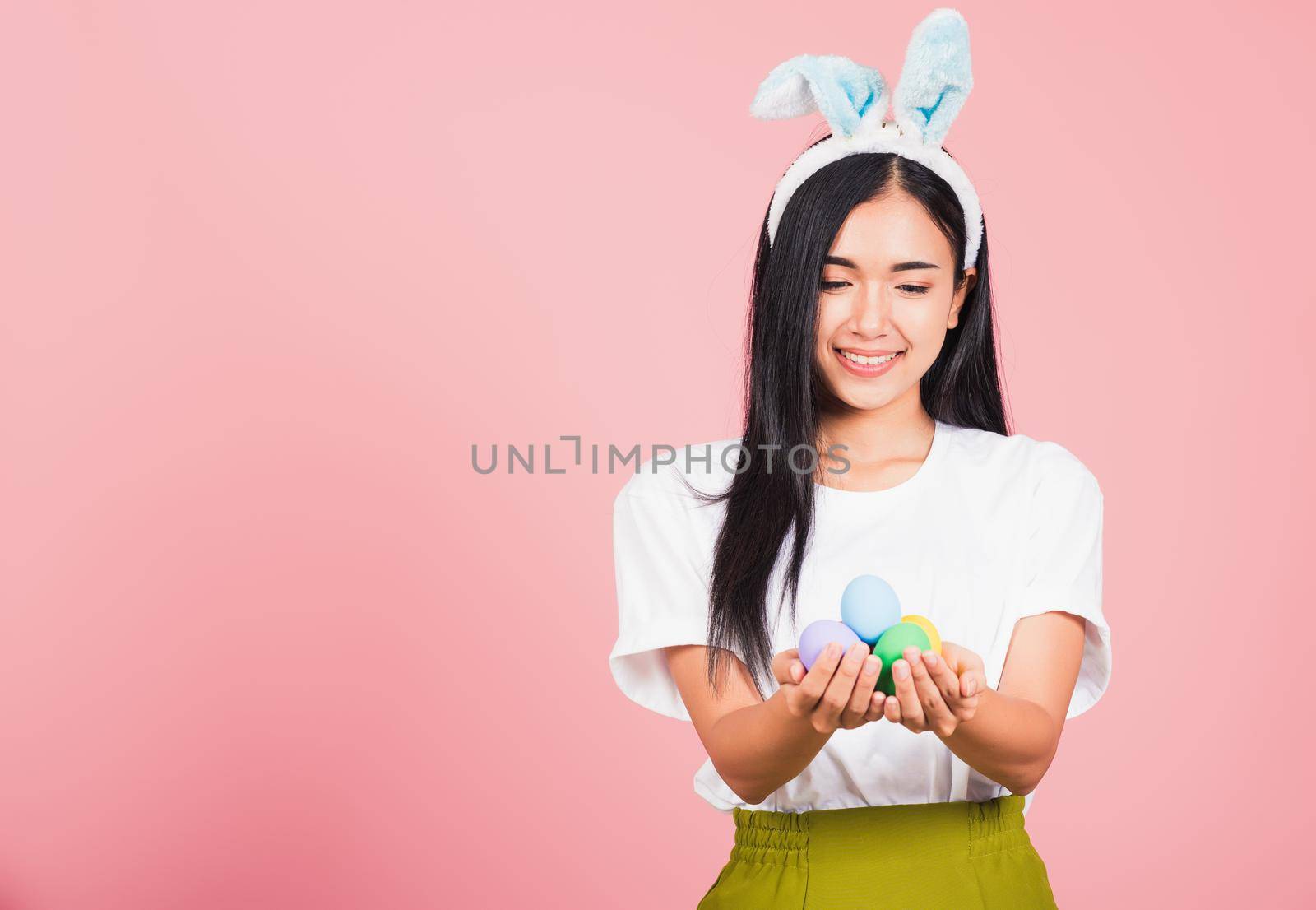 Happy Easter concept. Beautiful young woman smiling wearing rabbit ears holding colorful Easter eggs gift on hands, Portrait female looking at eggs, studio shot isolated on pink background
