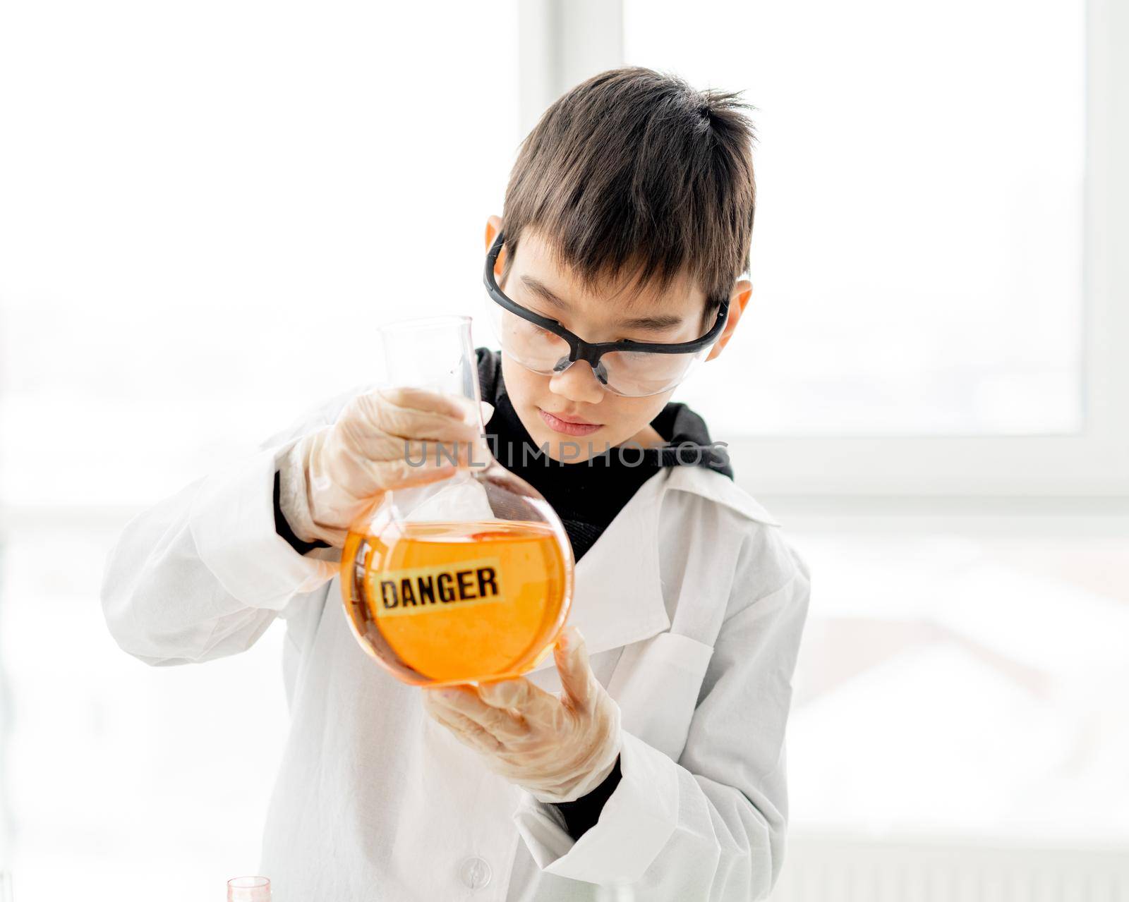 School boy wearing protection glasses doing chemistry experiment in elementary science class. Clever pupil holding tube with danger mark in lab during test