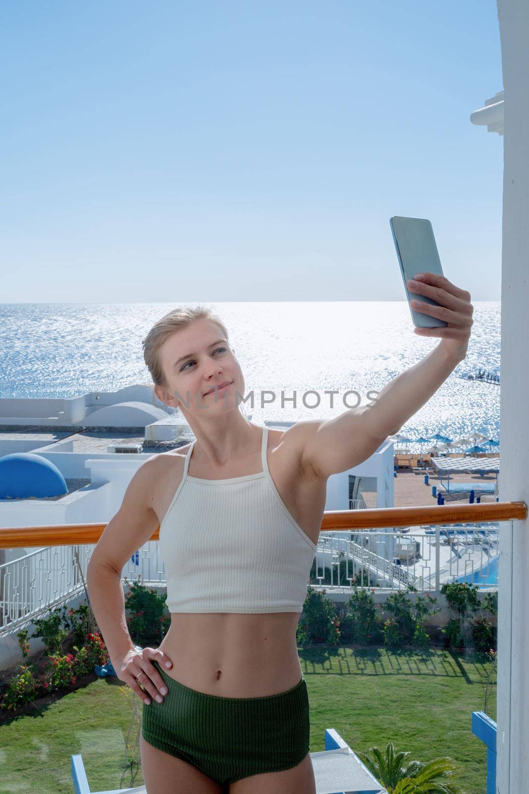 Vacation and travel. Happy woman in swimsuit taking selfie on mobile phone standing on the balcony of the hotel room, sea view