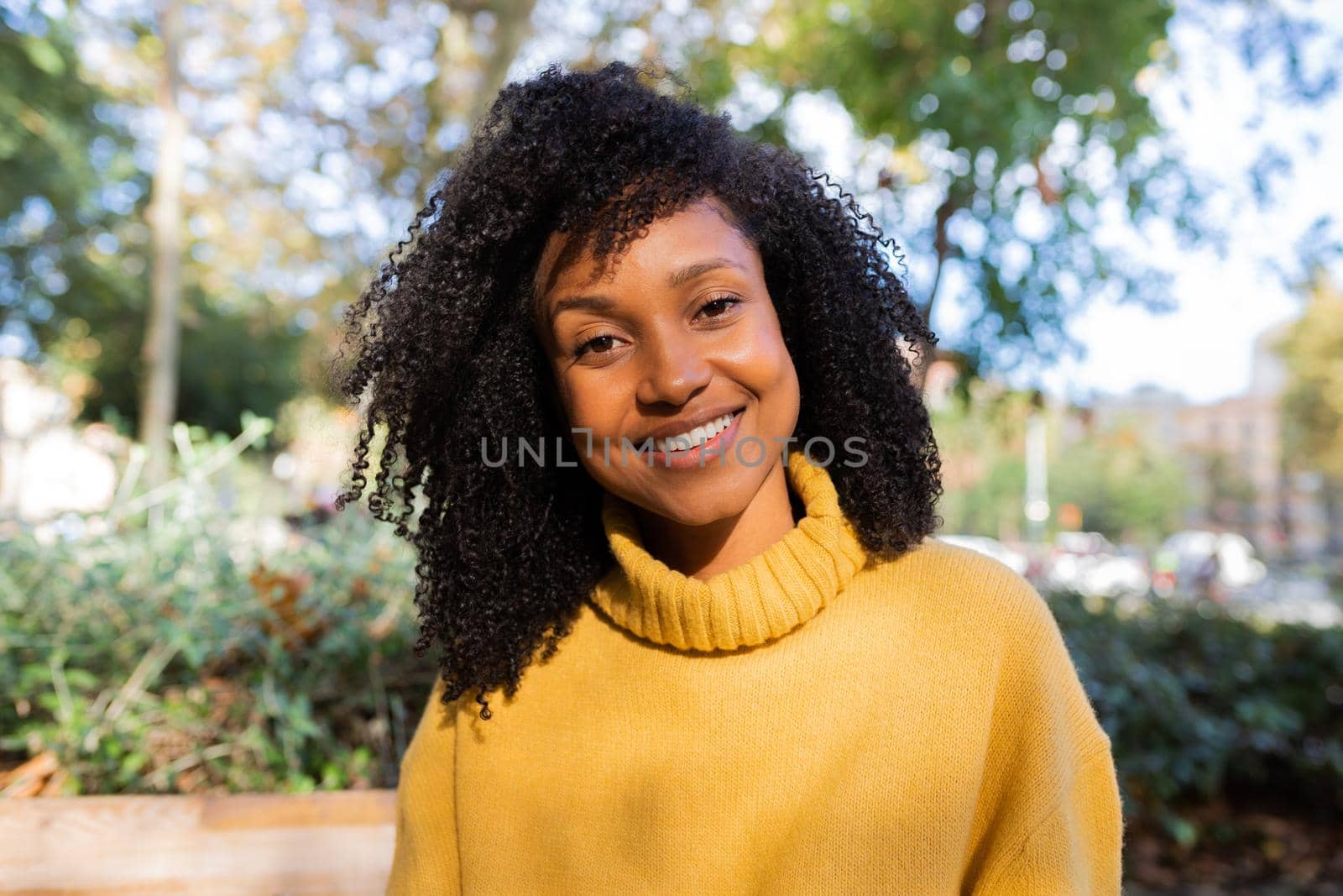 Portrait of smiling African American woman looking at camera wearing yellow sweater in a park. by Hoverstock
