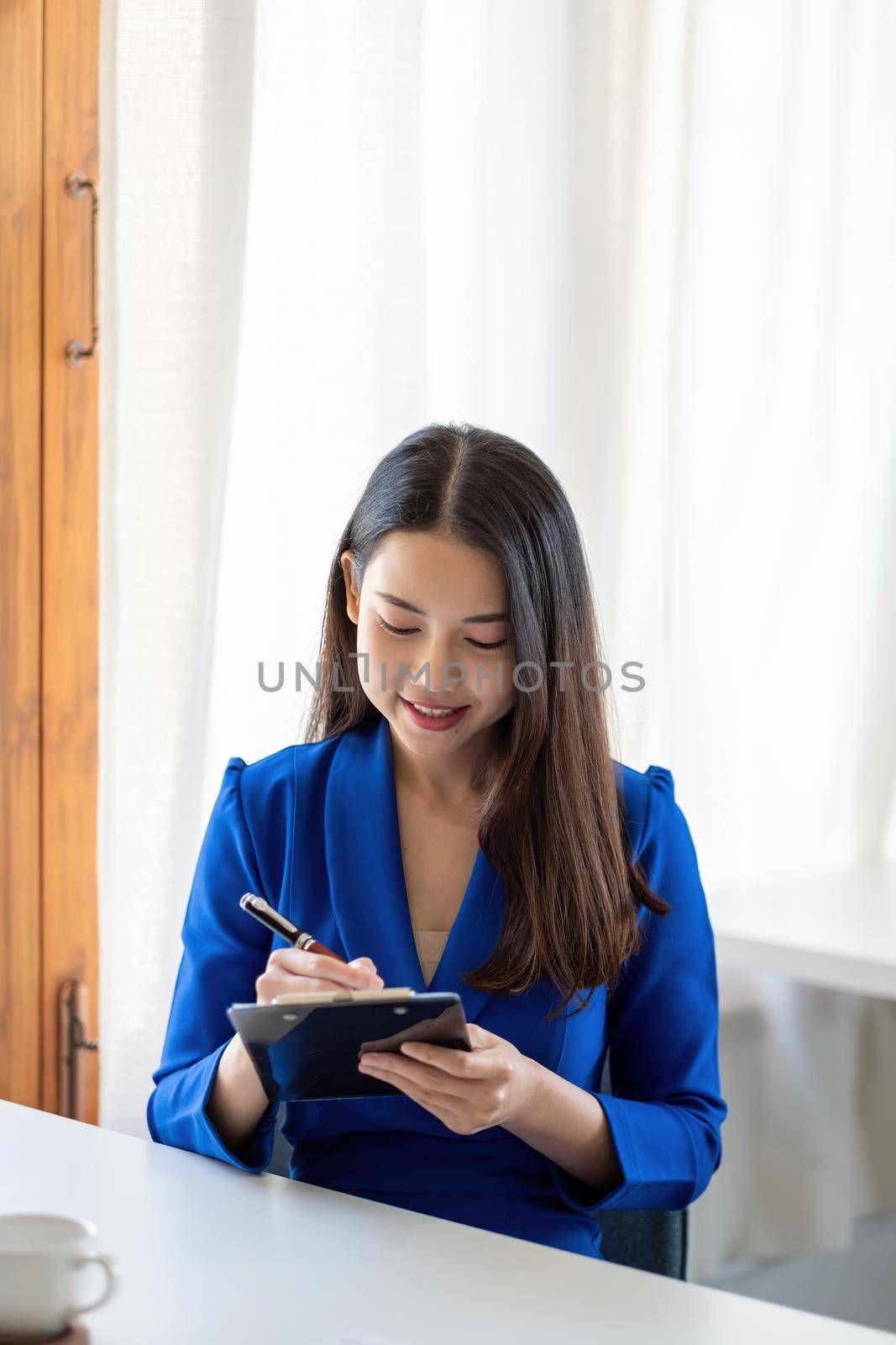 Portrait of a happy asian businesswoman using pen for taking note on notepad at home office.
