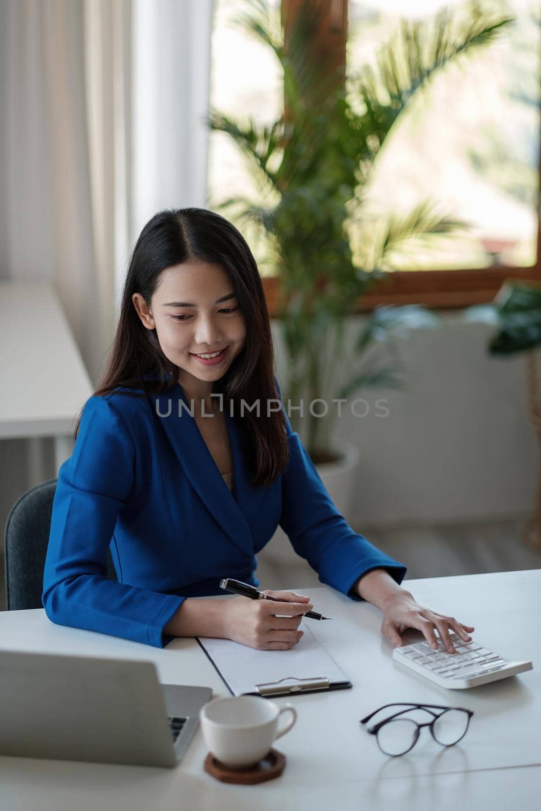 Portrait young asian businesswoman sitting at their desk and take notes using laptop and calculator in the office. by nateemee