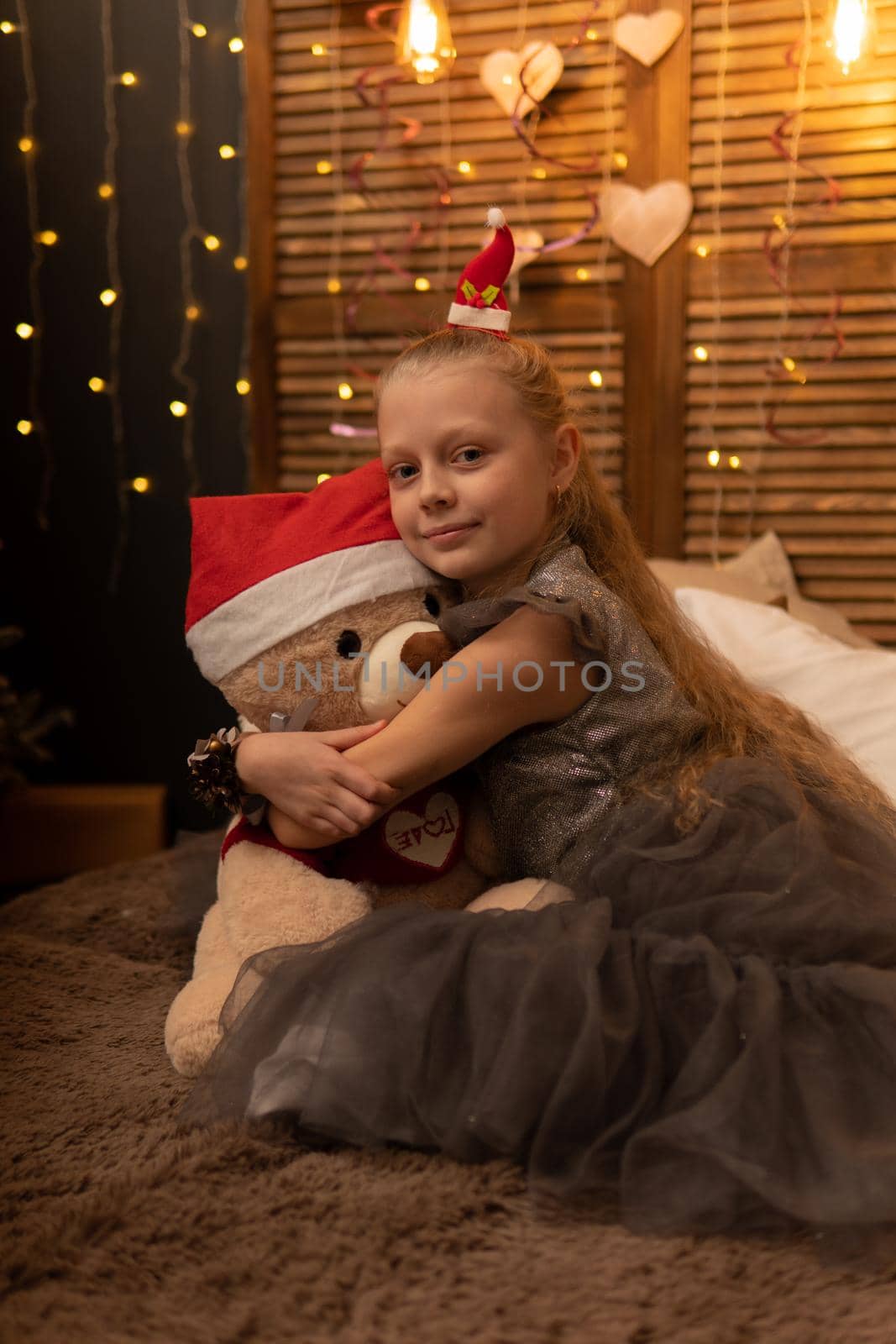 A girl hugs a bear toy in a Christmas cap bear Christmas kid girl cute, from people young in comfort sitting room, adorable joyful. Bedroom woman holding, hug wake the concept of kindness