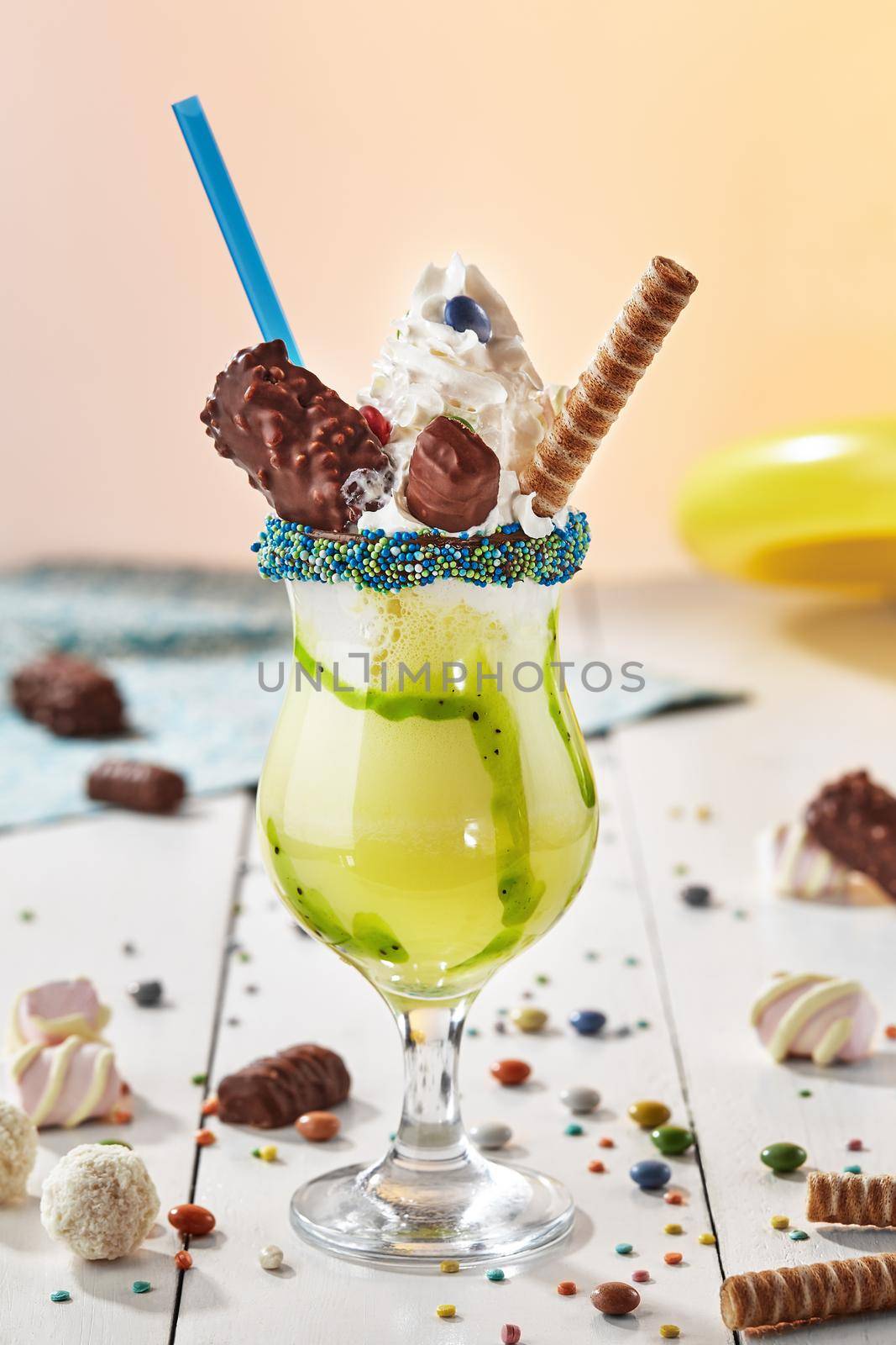 Exotic fruit milkshake with kiwi topped with whipped cream, waffle roll, candy bars, marshmallow, coconut balls and colorful dragees in glass garnished with sprinkles. Children party menu concept