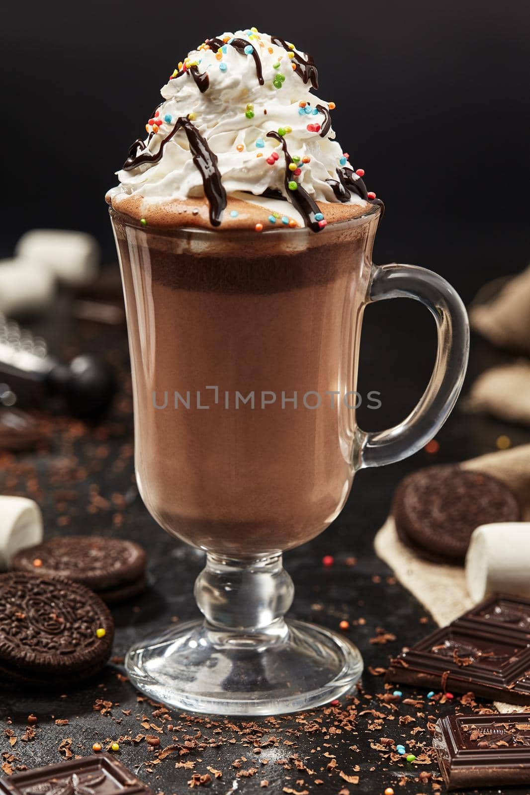 Delicious aromatic hot chocolate with whipped cream topped with colorful sprinkles in glass cup on black table with cookies, pieces of chocolate and marshmallows