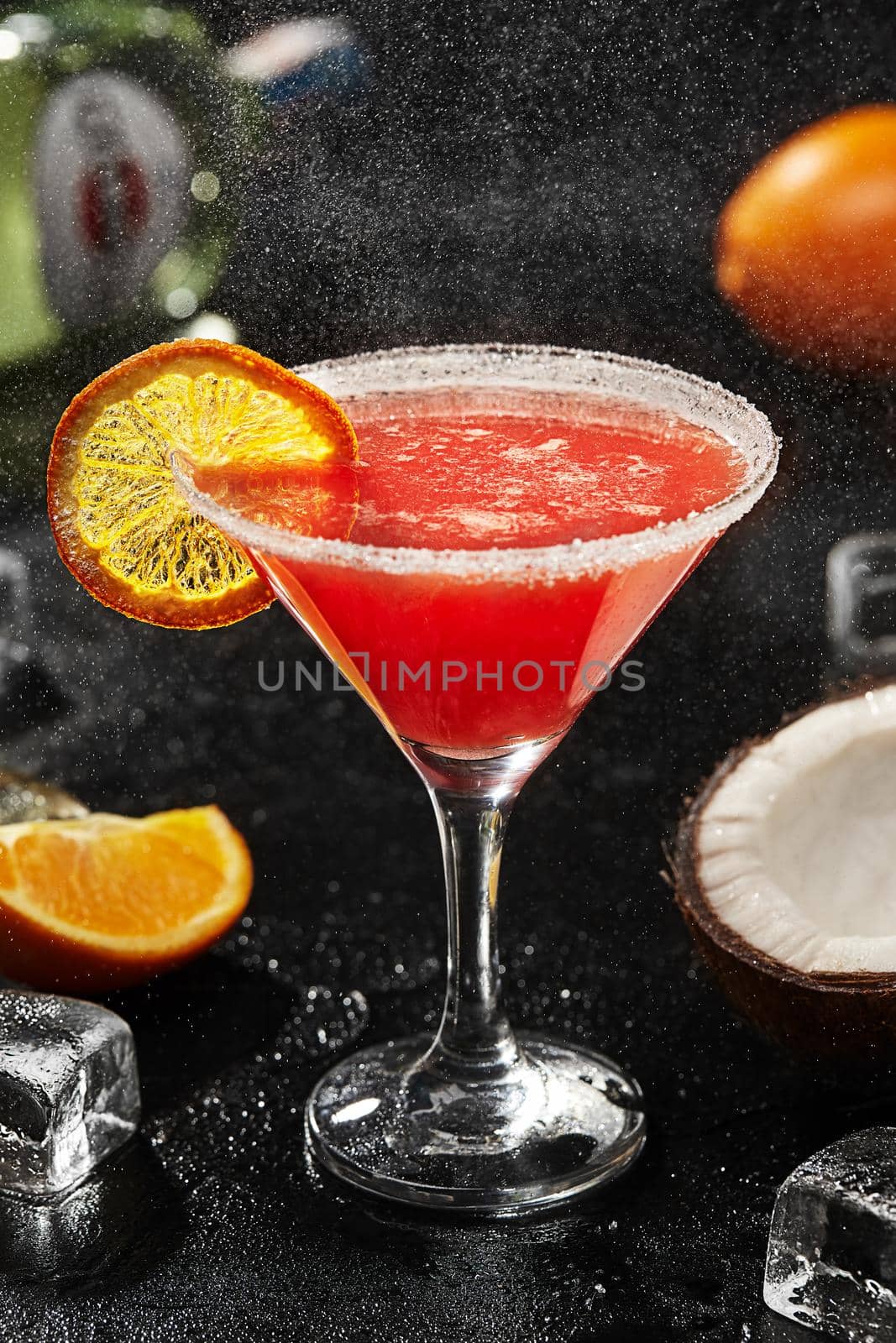 Exotic cocktail from martini with tropical flavours of coconut liqueur, citrus notes of orange juice and sweetness of pomegranate syrup garnished with candied orange slice on black background