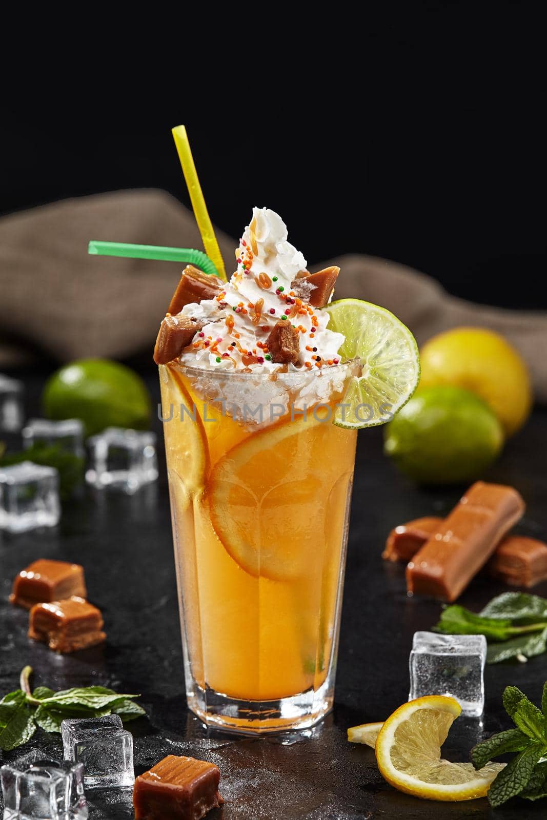 Delicious refreshing lemonade with lime and orange slices topped with delicate whipped cream, sweet toffees, caramel sauce and colorful sprinkles on black background. Unusual mix of drink and dessert