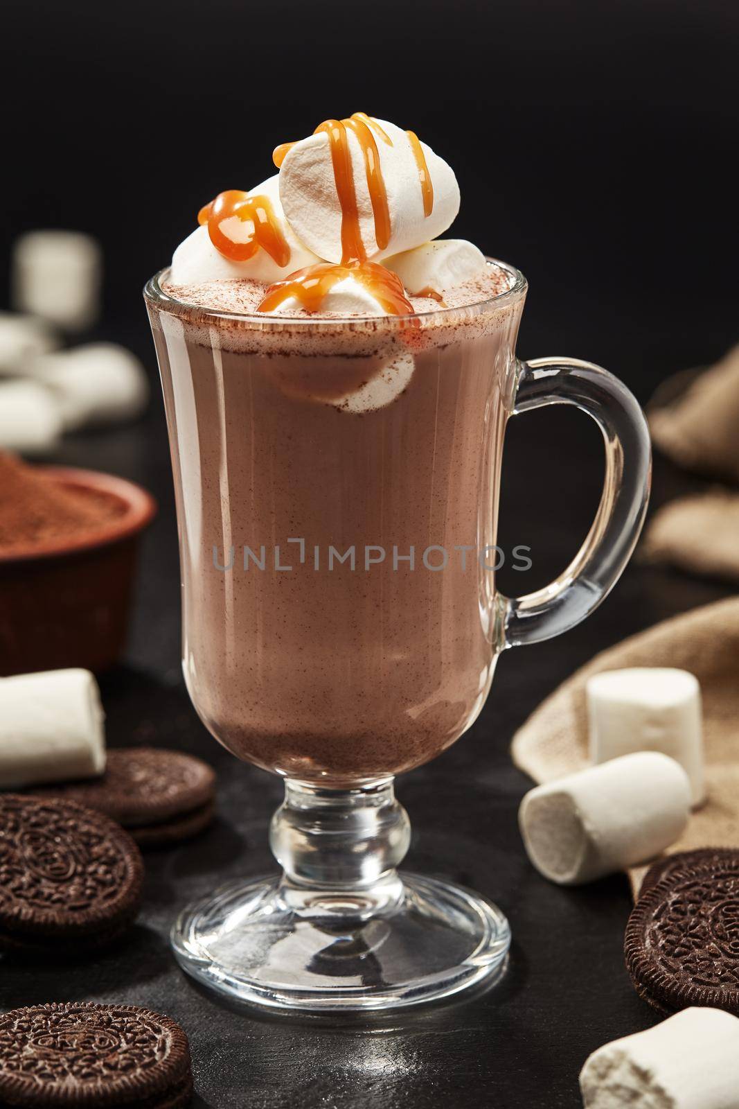 Glass cup of hot cocoa with marshmallows and caramel topping on black table with chocolate cookies and cocoa powder in bowl on background, selective focus