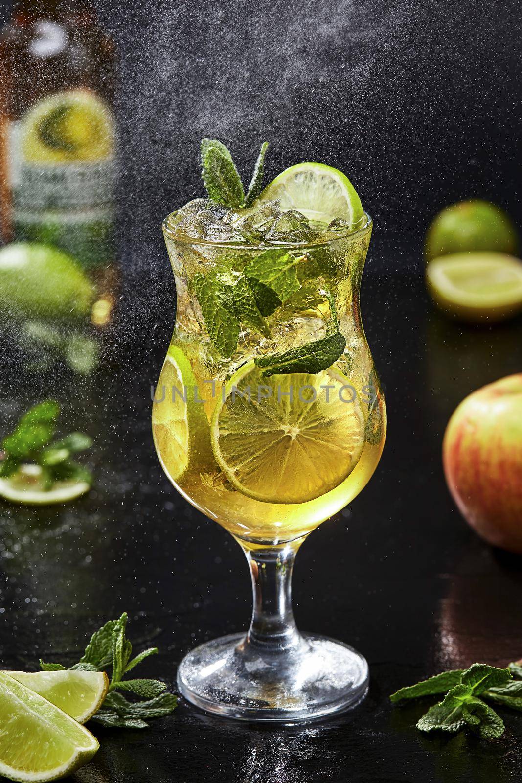 Fresh low alcohol cocktail from apple cider with lime slices and fragrant mint leaves served with crushed ice in glass on black background with ingredients in fine water spray